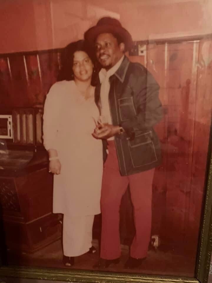 Couple posing together in a vintage indoor photo: woman in a light dress and man in a hat, denim jacket, and red pants.