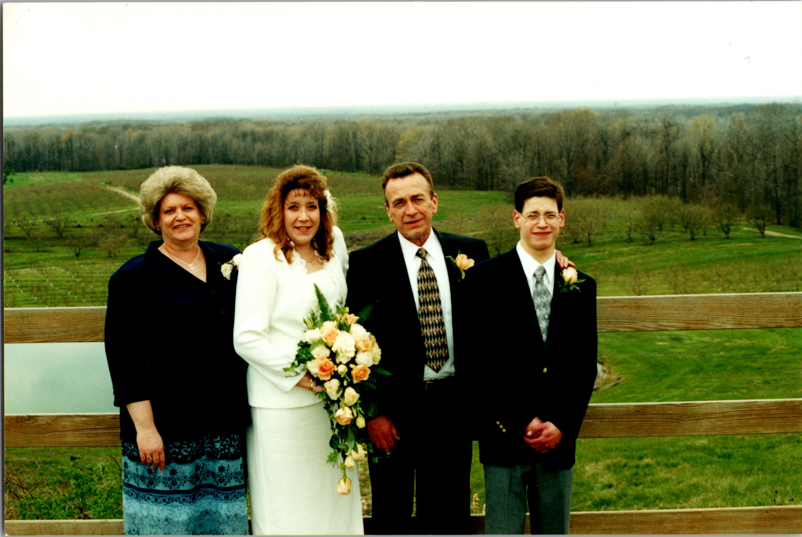 Bride in white holding a bouquet flanked by two men in suits and a woman in a patterned dress outdoors on a wooden railing with a countryside backdrop.