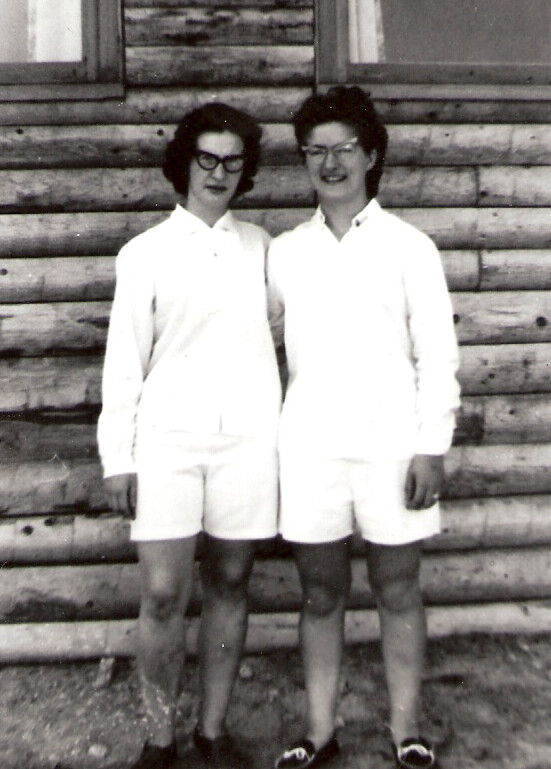 Two women in white shirts and shorts stand side by side in front of a log cabin wall.