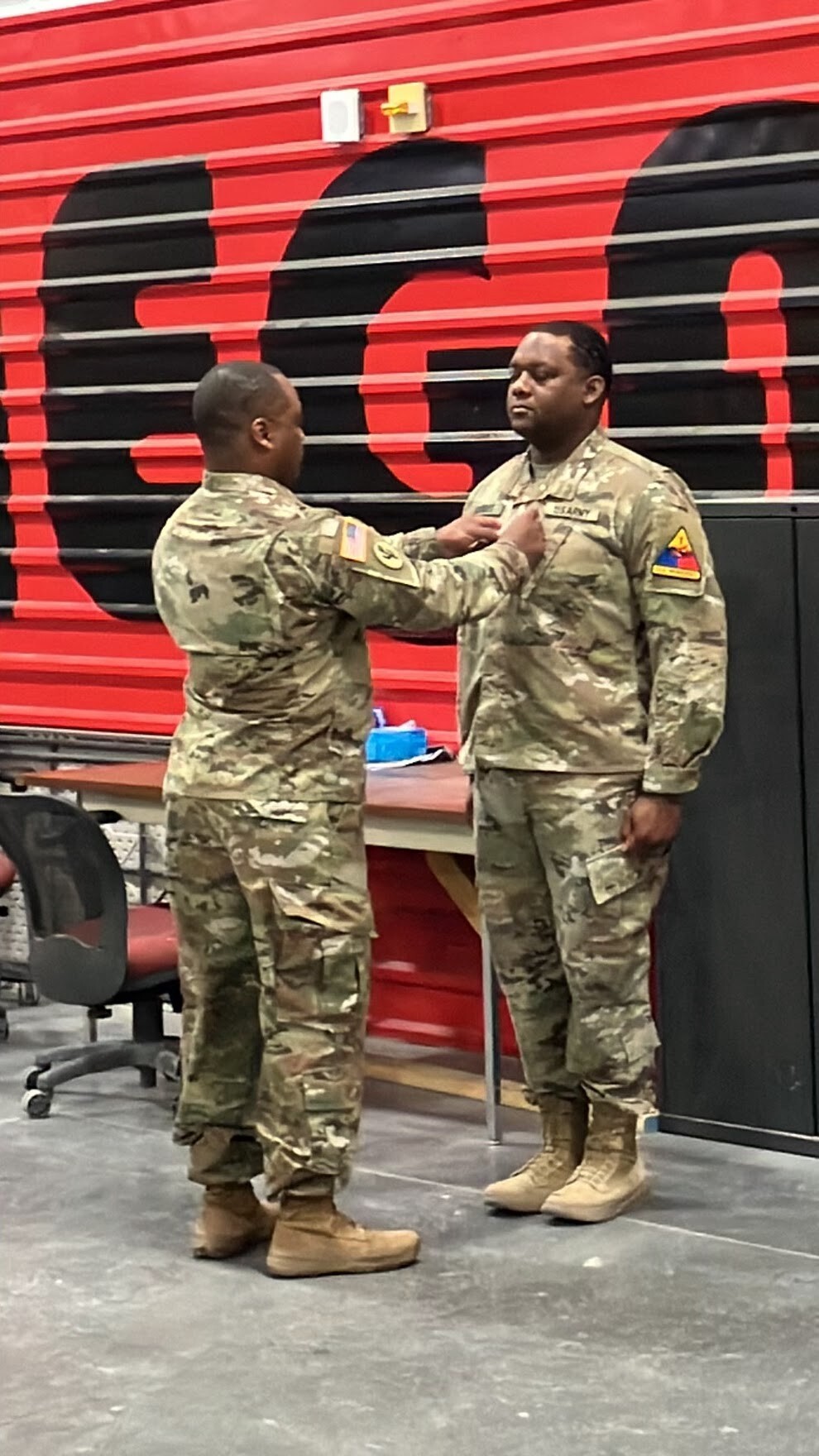 Two soldiers in camouflage uniforms pin a rank insignia on a comrade's chest during a ceremony in a gym with a red wall backdrop.