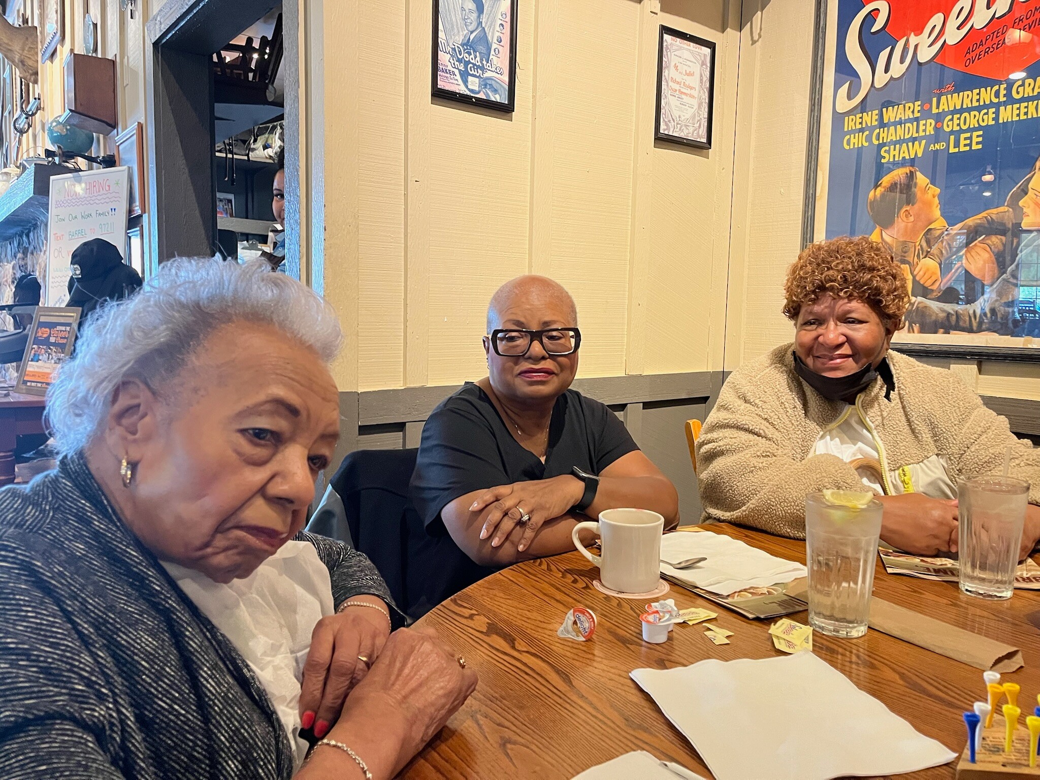 Three women sit at a wooden table in a cafe, smiling, with cups, glasses and napkins on the table in front of them.