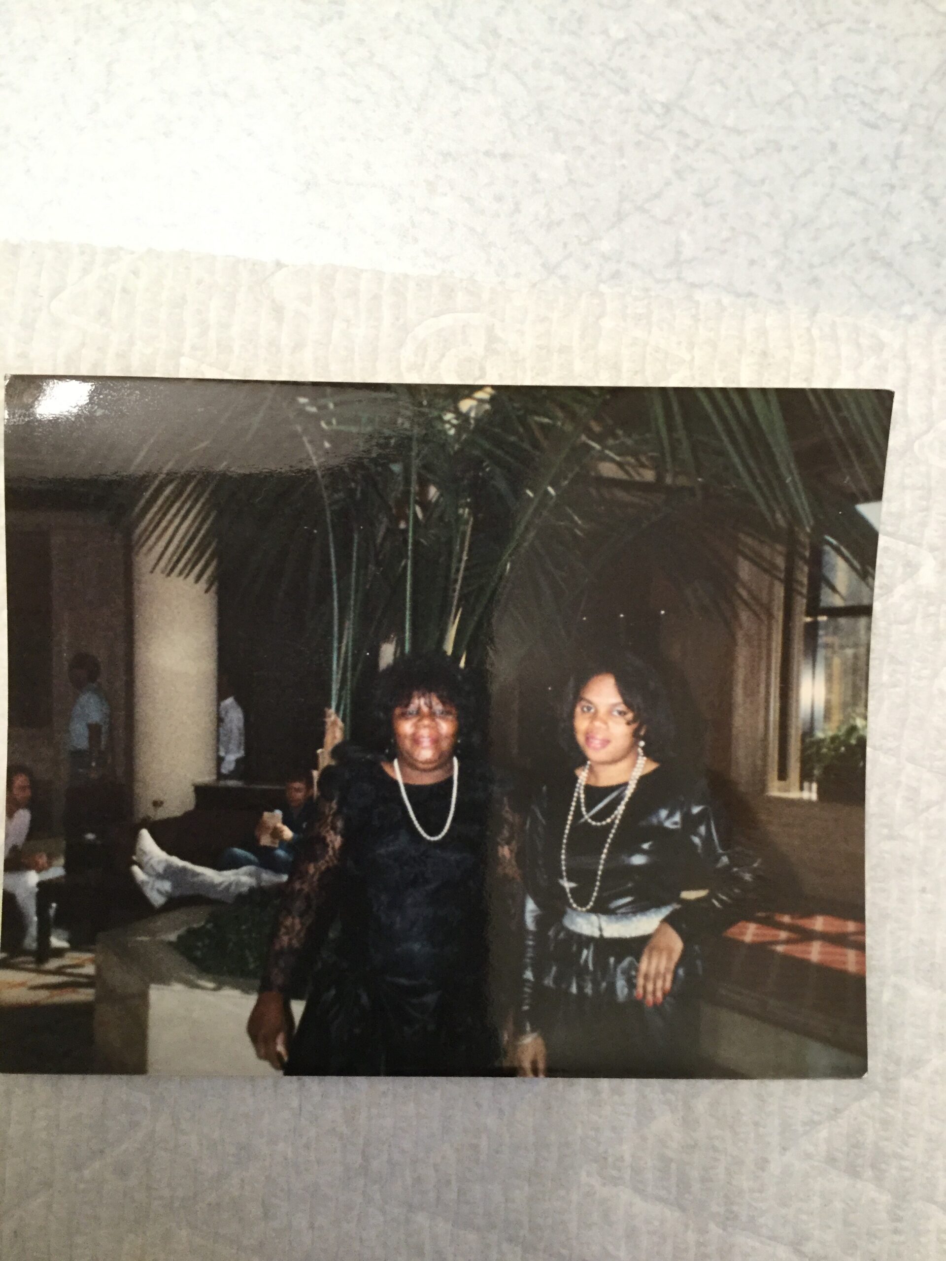 Two women in formal black dresses with pearl necklaces posing indoors, plant decor behind them.