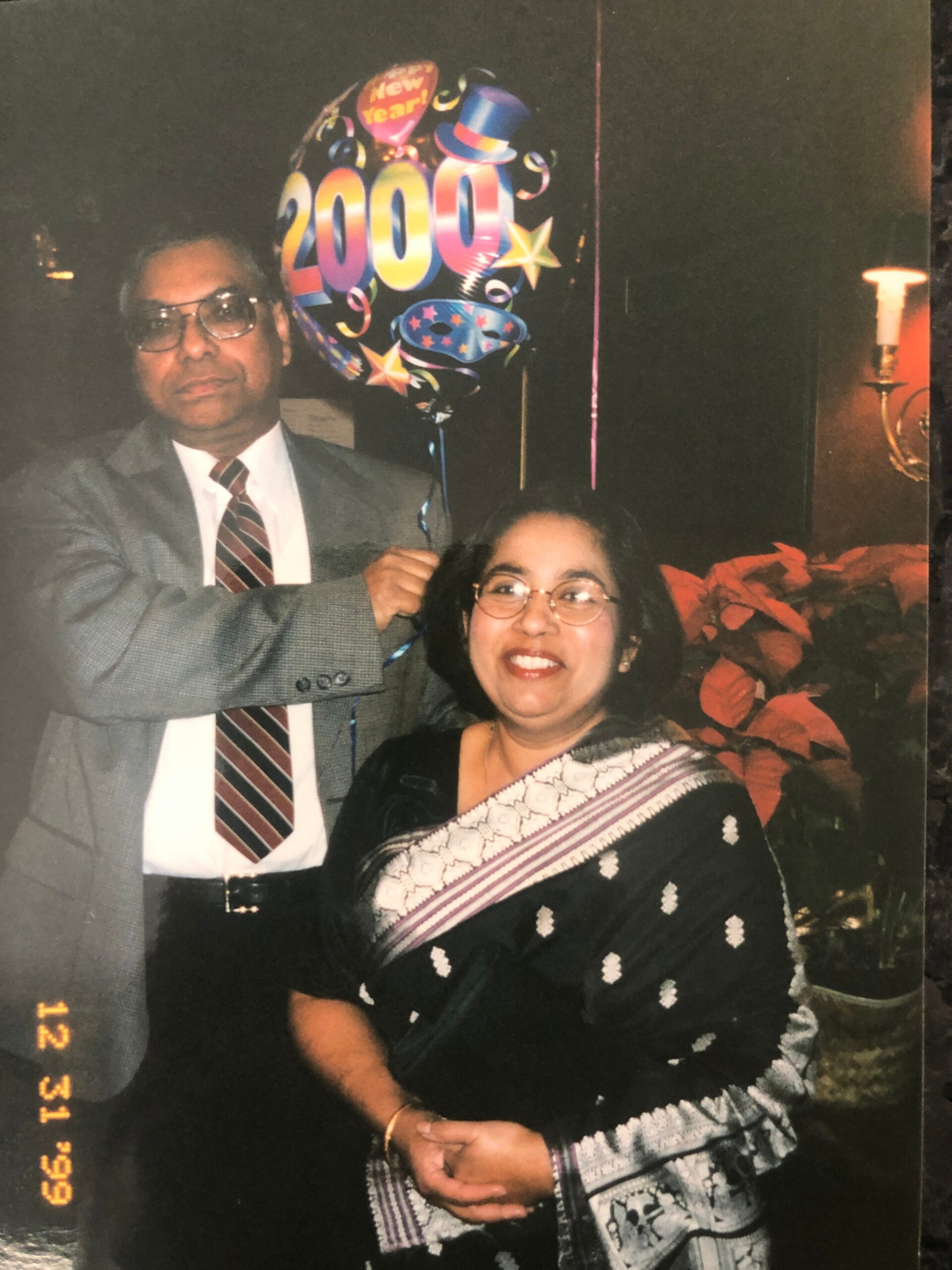 Man in a suit and tie stands with a smiling woman in a sari at a New Year party, holding a colorful 2000 balloon beside them.