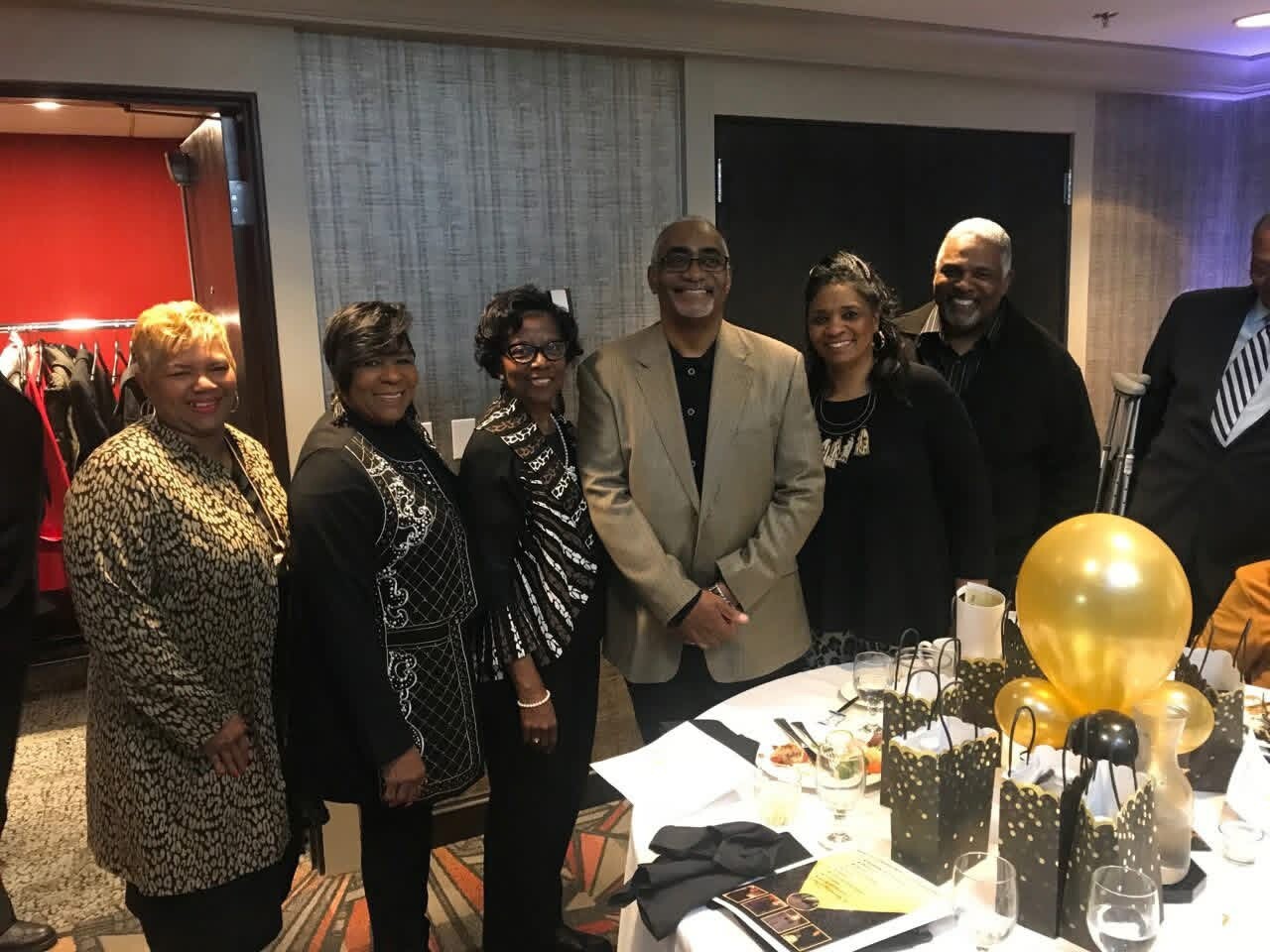Group of six adults smiling for a photo around a round table with black-and-gold gift bags and balloons in a banquet room.