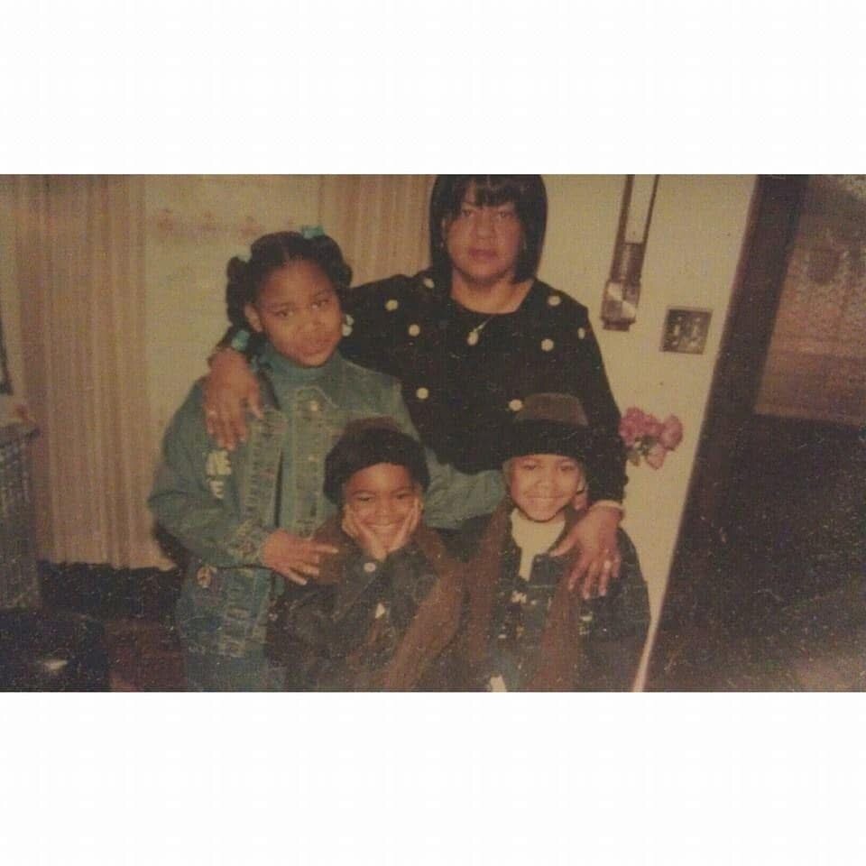 Four siblings pose for a casual indoor photo: two girls in denim jackets standing behind, a boy in front making a peace sign, and a younger child wearing a hat on the right, all smiling