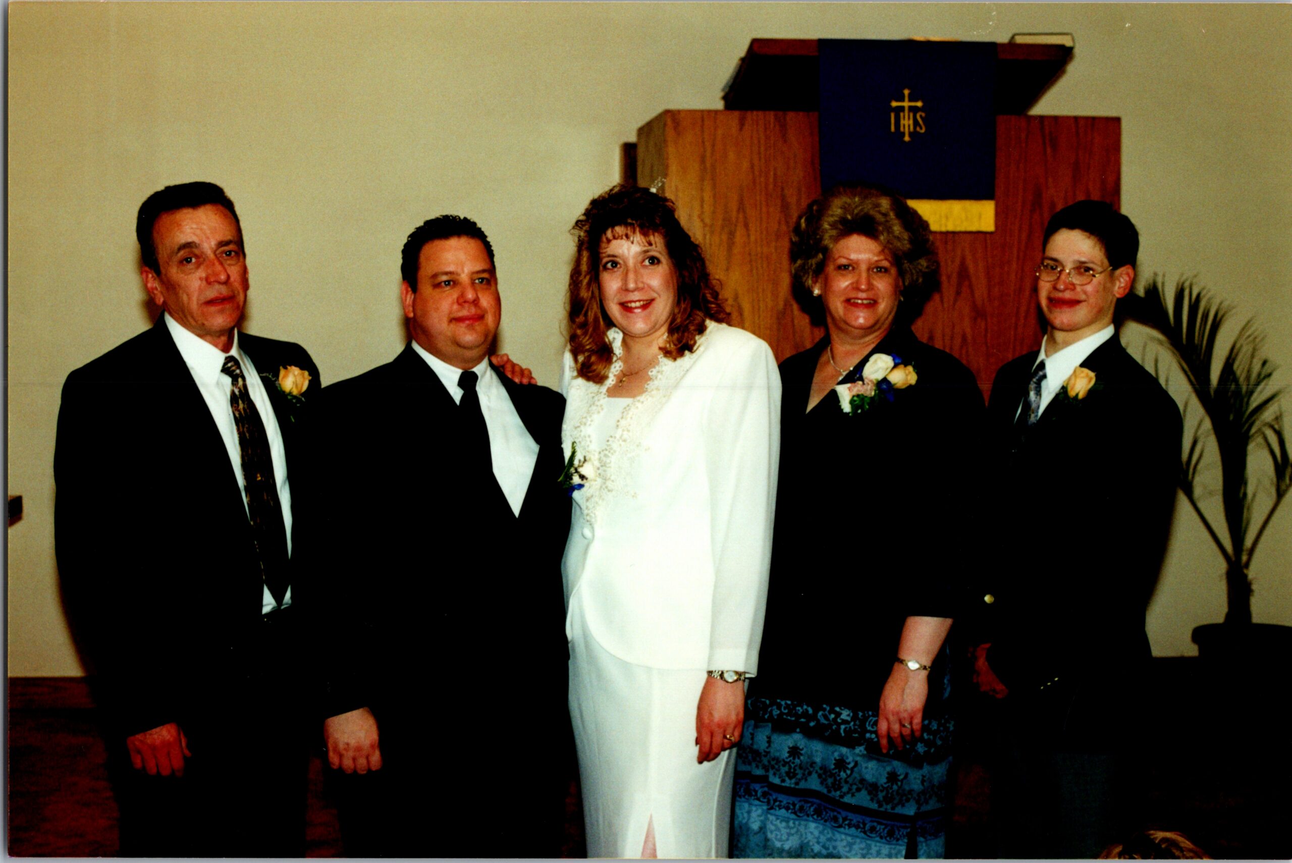 Bride in a white wedding gown flanked by four adults in formal wear, posing indoors with a church altar backdrop and floral boutonnieres.