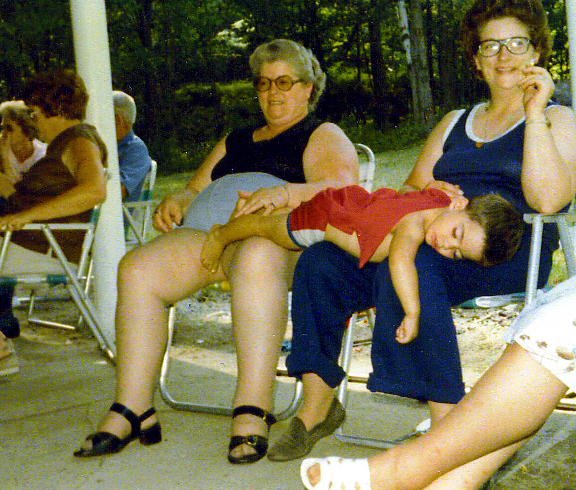 Two women sit on folding chairs outdoors; a sleeping child in a red outfit rests across the lap of the woman in black, while the woman in blue smiles nearby.