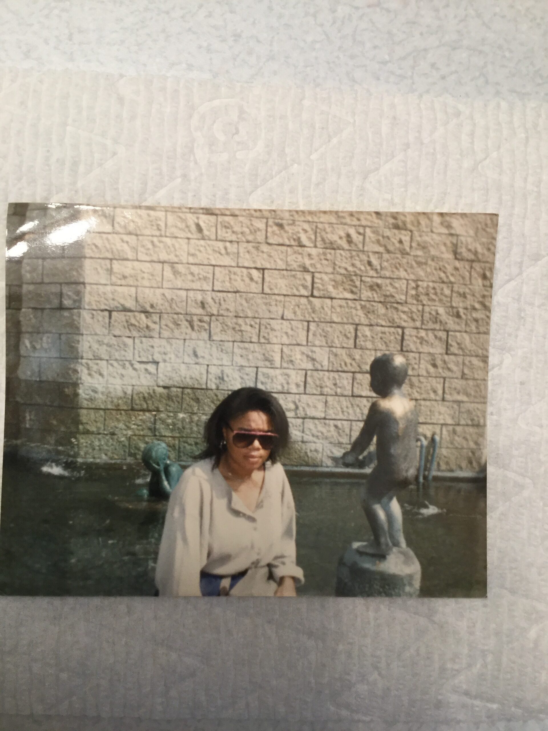 Woman wearing sunglasses sits by a shallow fountain with a stone statue in the background, in front of a brick wall backdrop (vintage photo).