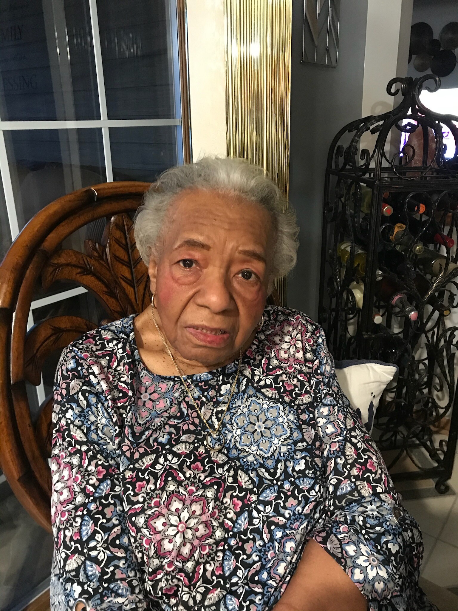 Portrait of an elderly woman with short gray hair wearing a colorful floral blouse, seated indoors by a decorative rack of wine bottles.