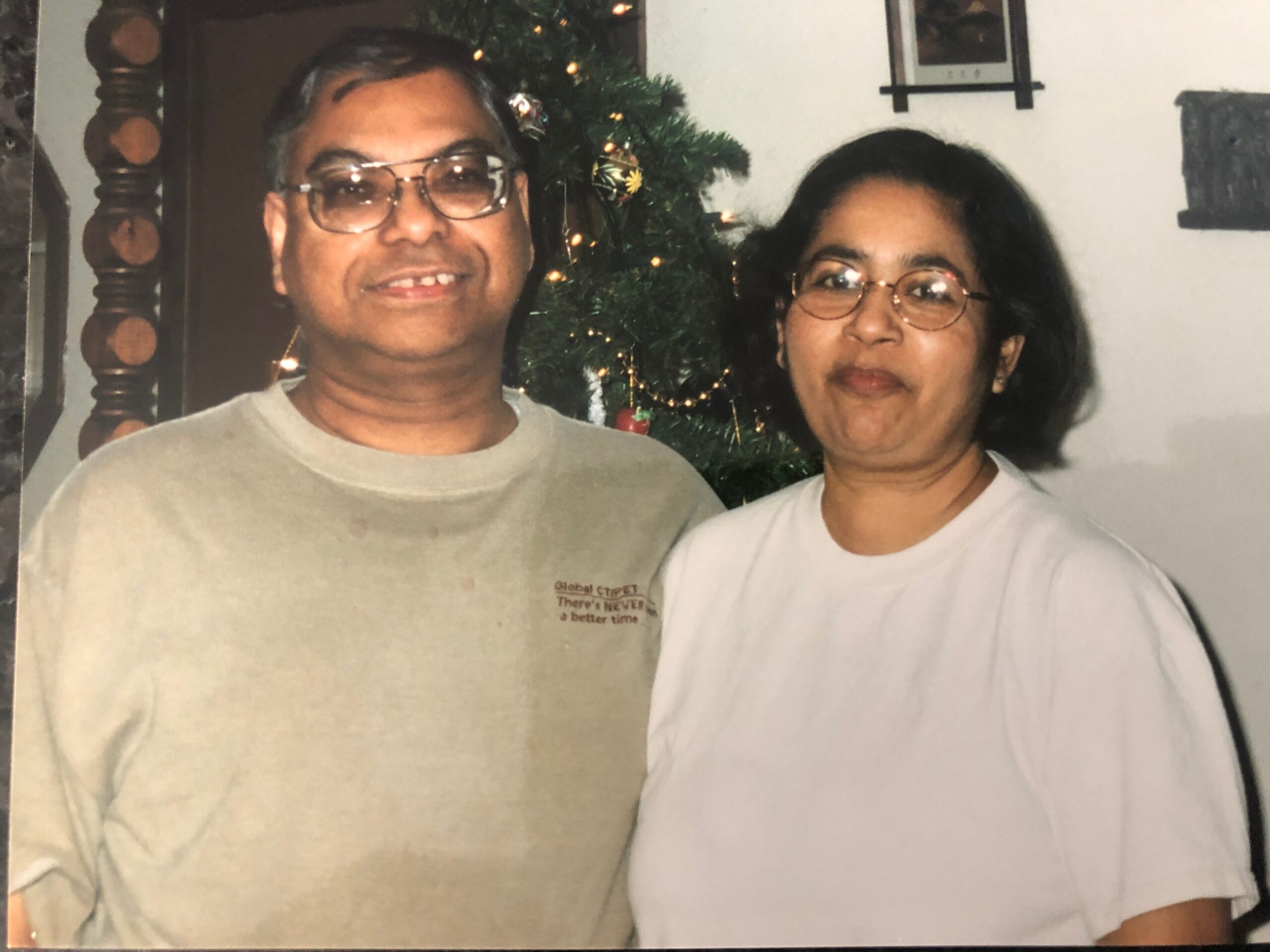 Smiling couple wearing glasses pose indoors, with a Christmas tree in the background behind them.