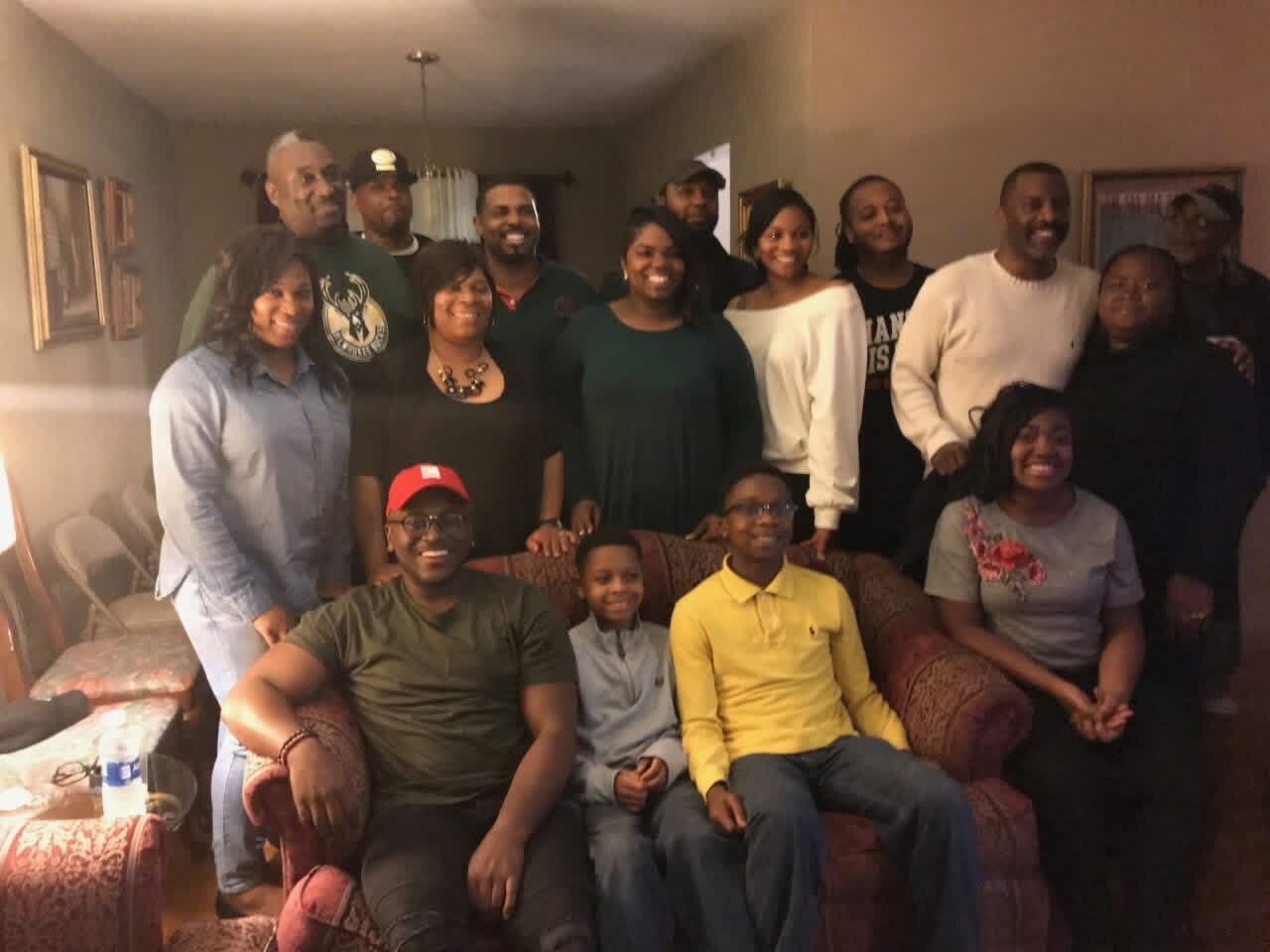 Large group of people posing for a photo in a living room, with two kids seated on a sofa and adults standing behind them.