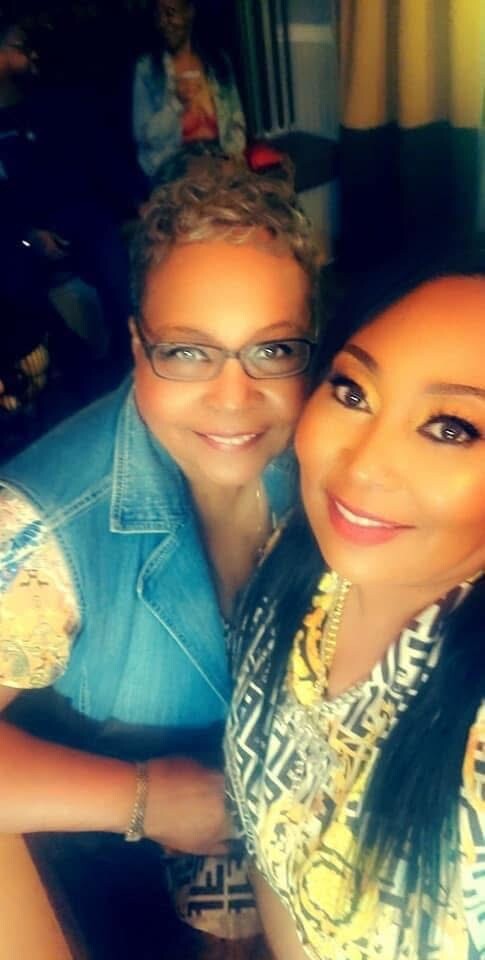 Two women pose for a selfie indoors, smiling at the camera; one wears glasses and a denim vest, the other is in a patterned blouse with bold makeup.