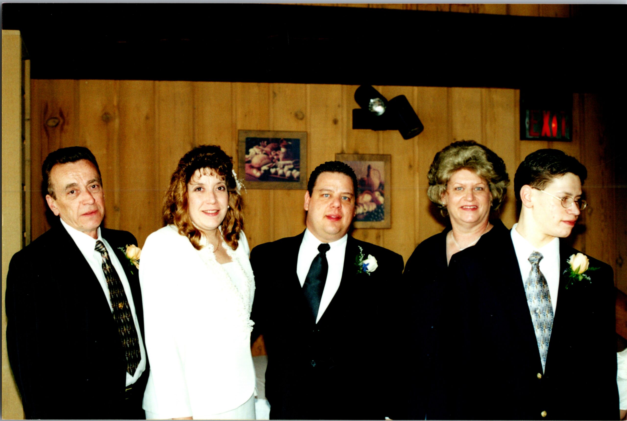 Bride in a white wedding dress standing with four guests in suits at a wedding reception, posing for a group photo against wood-paneled walls and visible exit sign in the background.