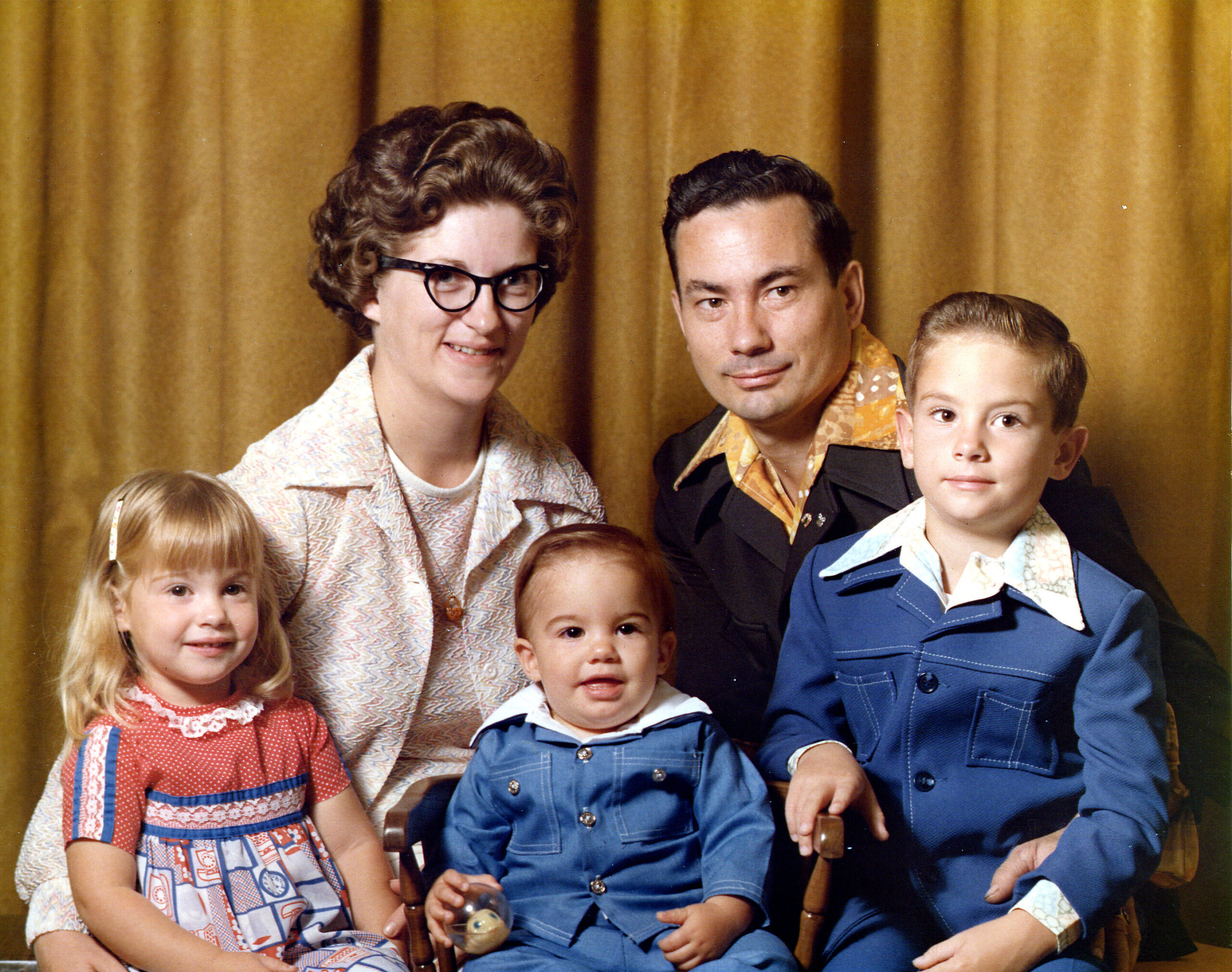 Group photo of a family posing in front of a gold curtain; children wearing blue overalls and adults in retro clothing with a woman wearing glasses in the center left and a man to her right.
