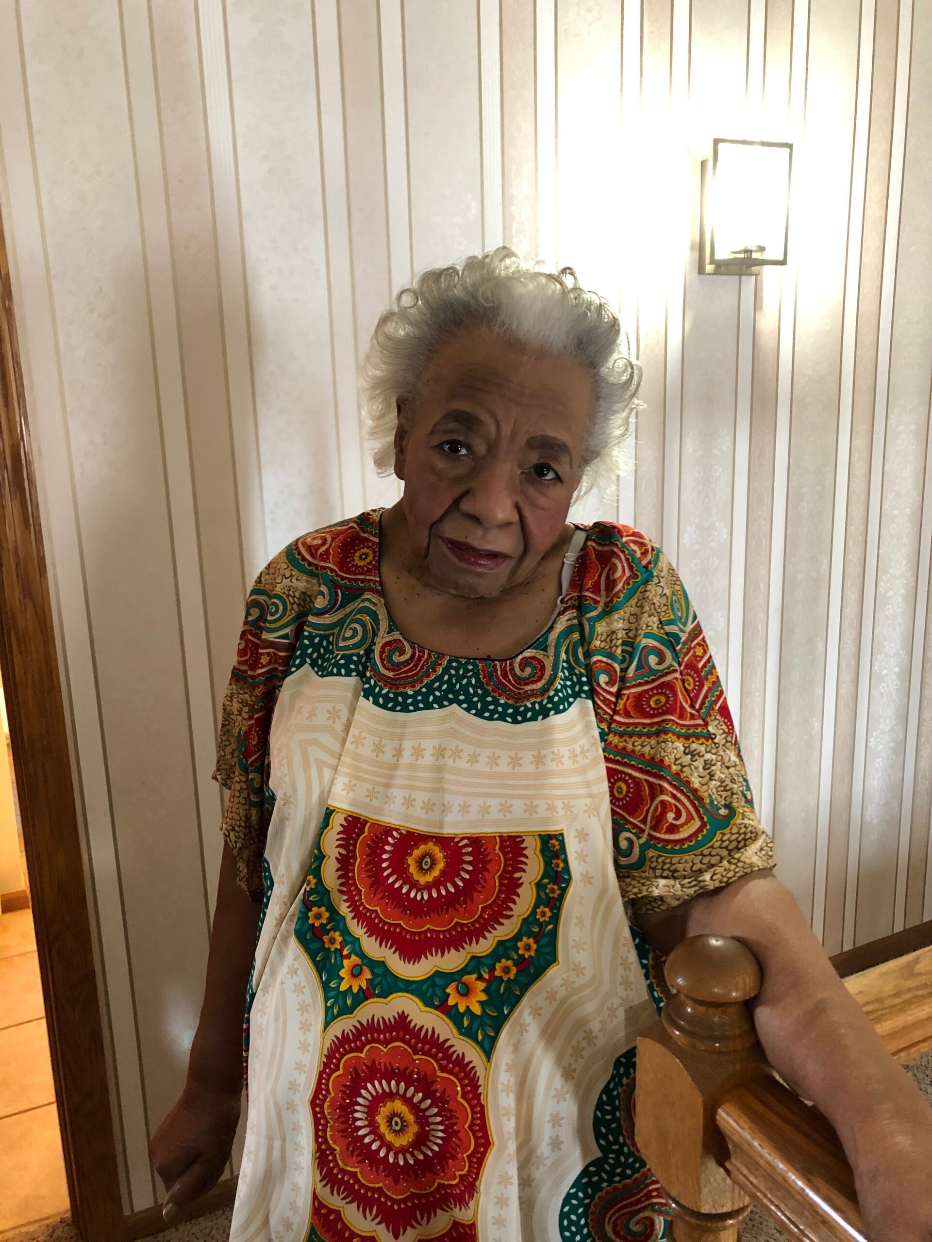 Elderly woman with white hair stands by a wooden railing in a patterned multicolor dress, looking at the camera.