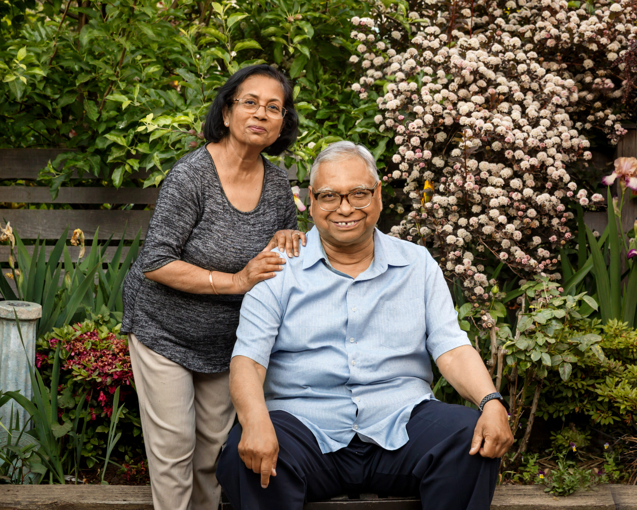 Two seniors pose together in a lush garden, the woman standing behind the seated man with her hand on his shoulder.