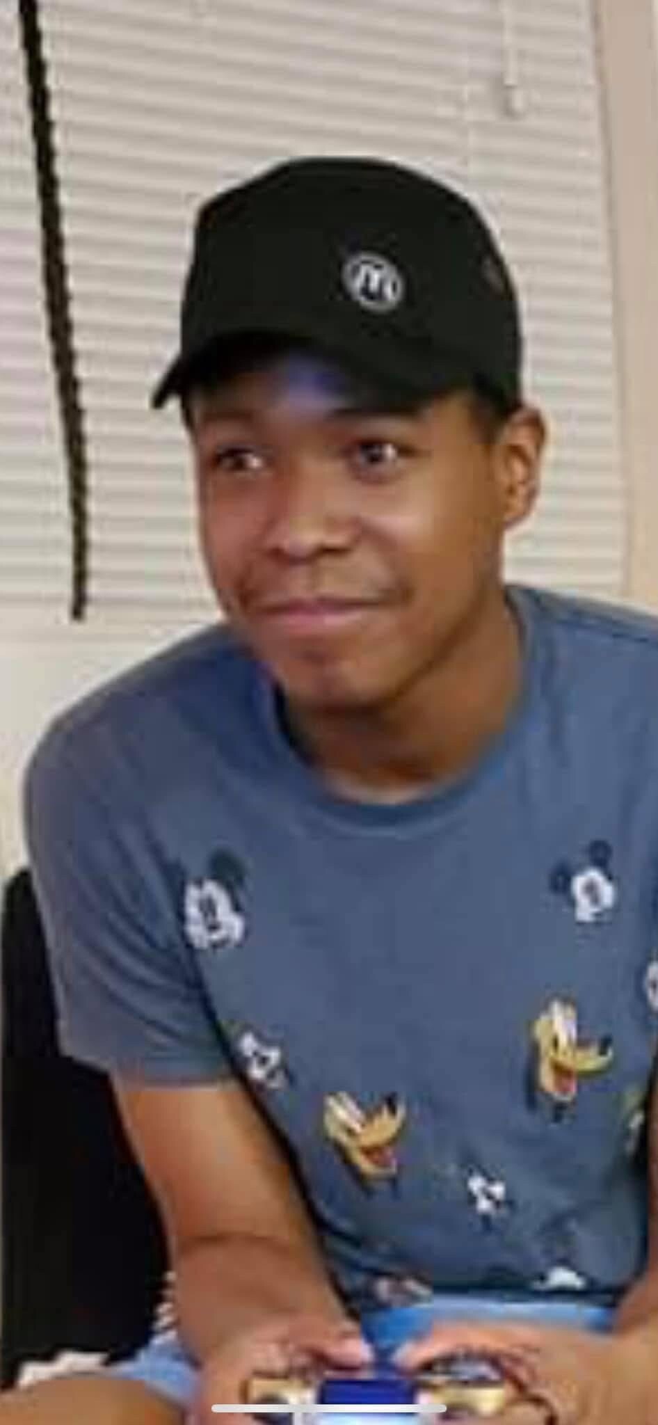 Young man wearing a black cap and blue T‑shirt, sitting at a table and holding a handheld device (phone/game).