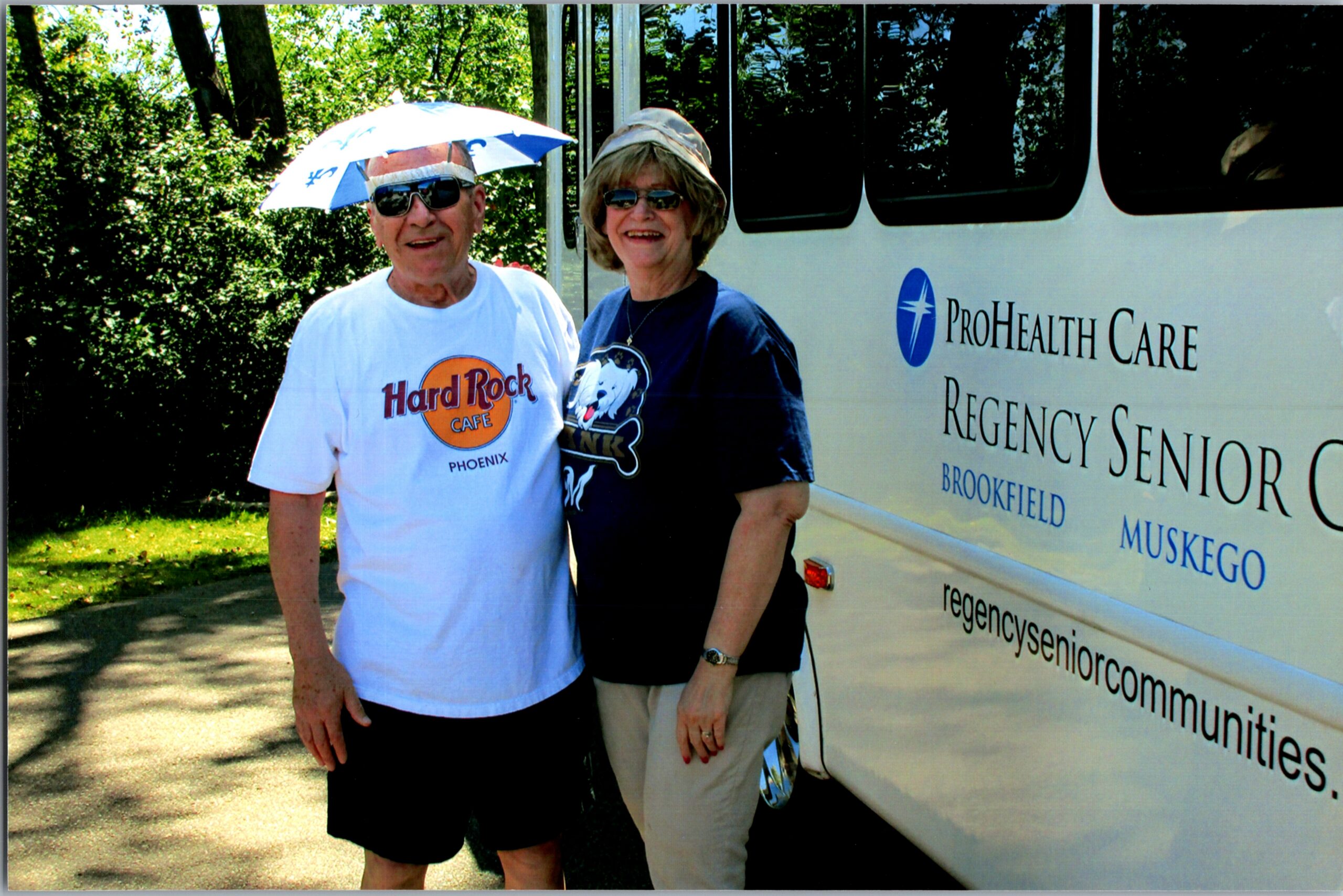 Two seniors wearing hats and sunglasses stand beside a ProHealth Care van outdoors, smiling at the camera (one in a Hard Rock Cafe Phoenix T-shirt).