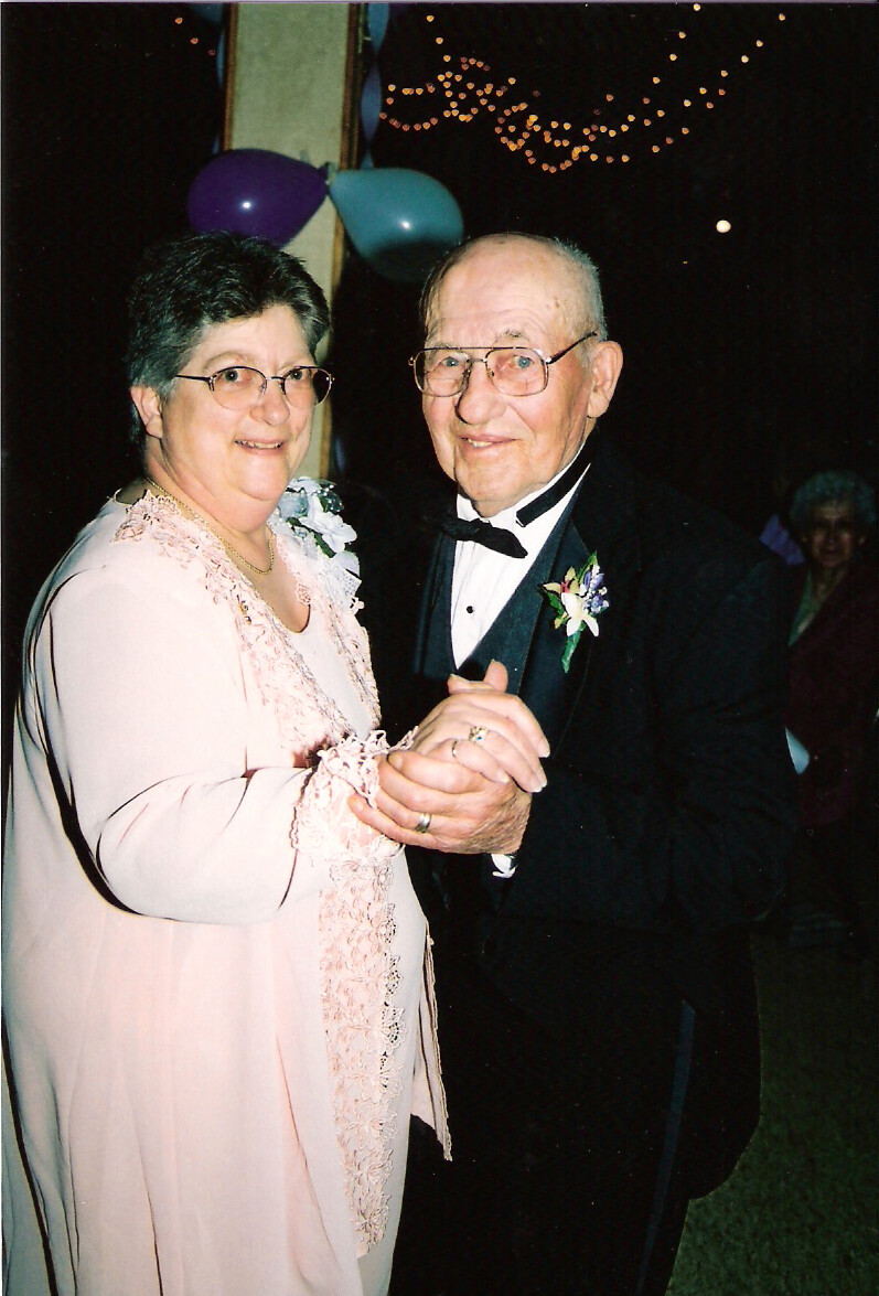 Older couple dancing together at a nighttime celebration; woman in pale pink lace dress, man in tuxedo with boutonniere, both smiling.