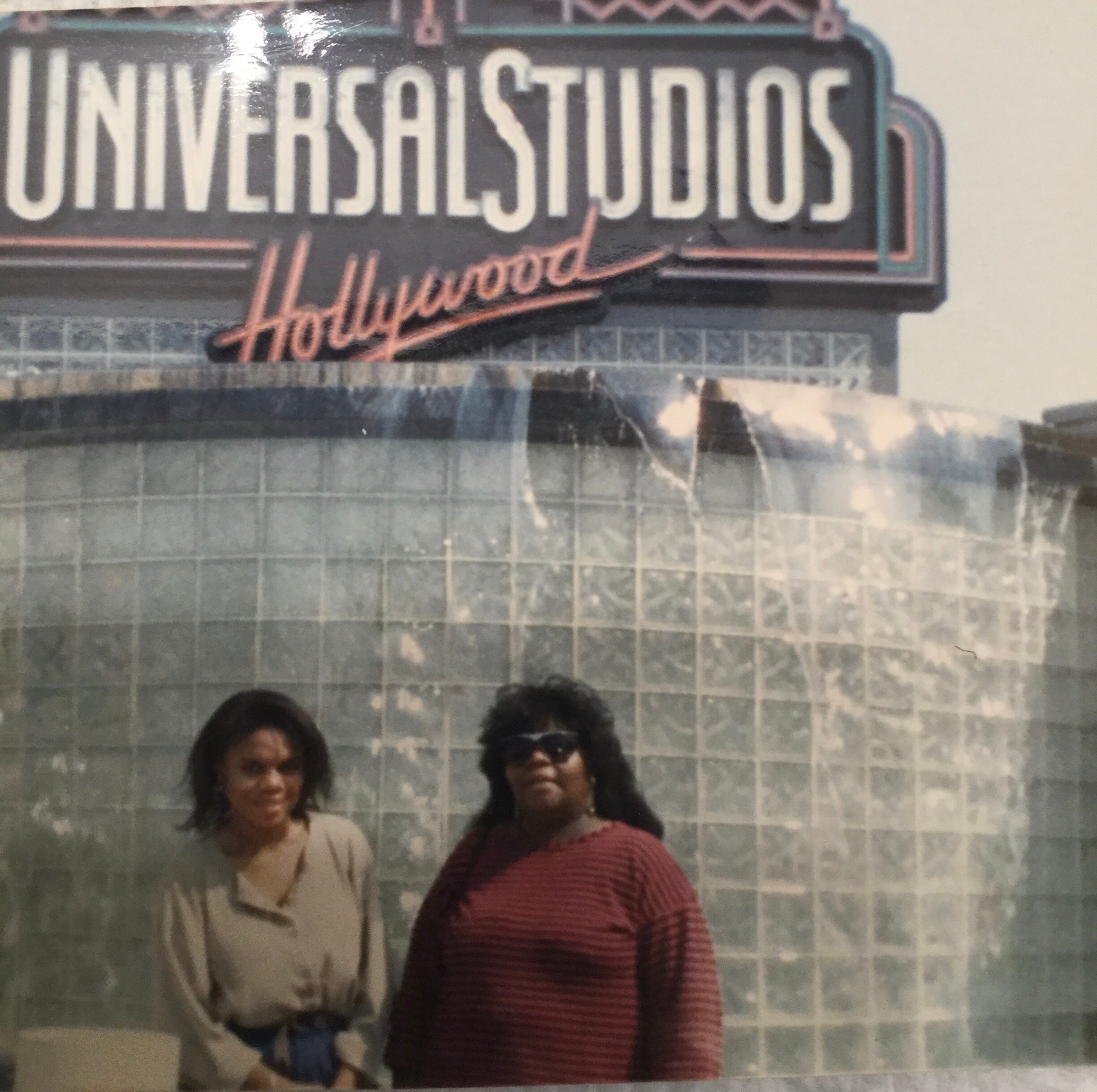 Two women pose in front of a tiled wall with a large vintage Universal Studios Hollywood sign graphic above them.
