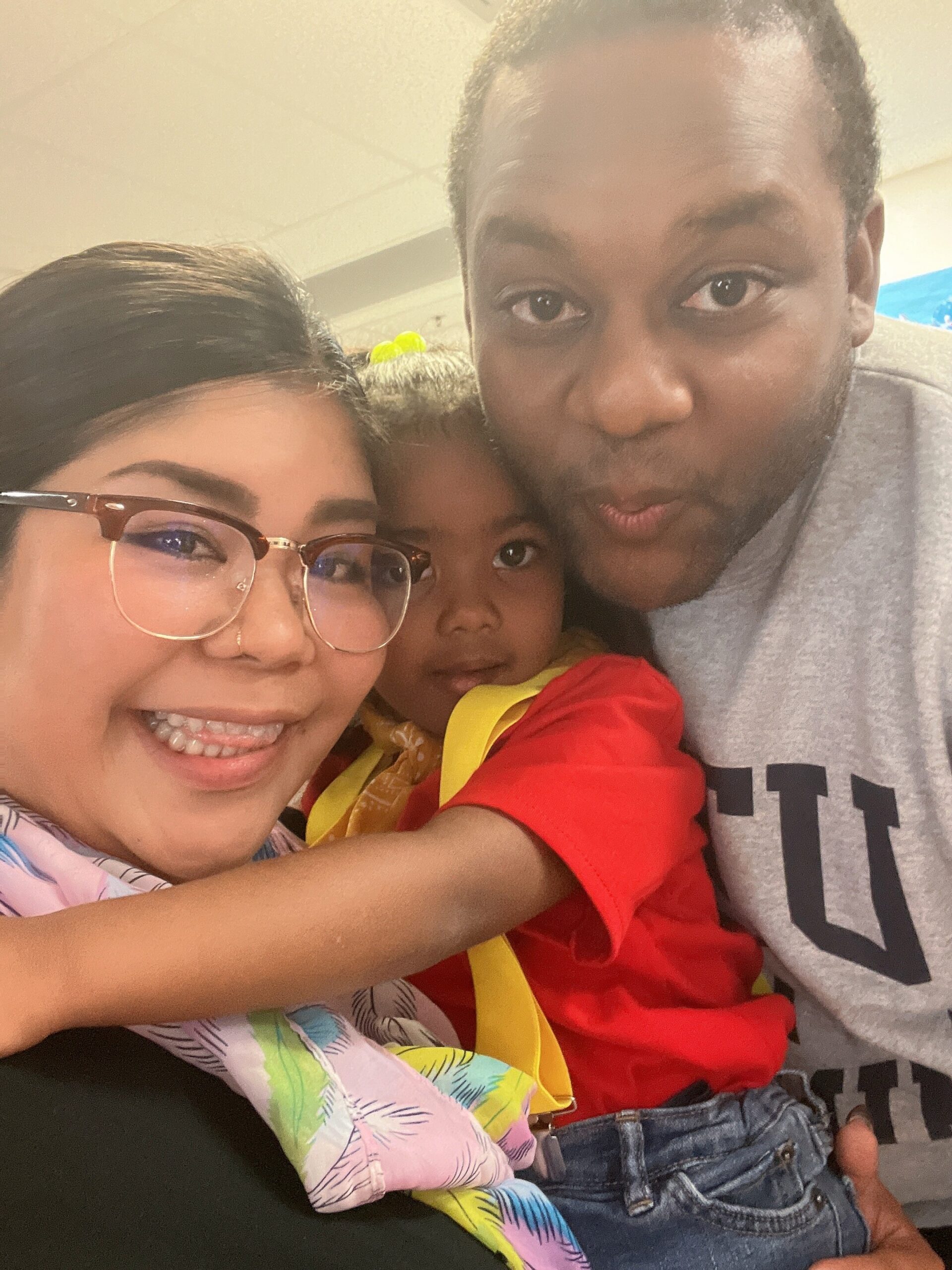 Close-up family selfie: a smiling woman with glasses on the left, a man kissing a young child wearing a red shirt in the middle.