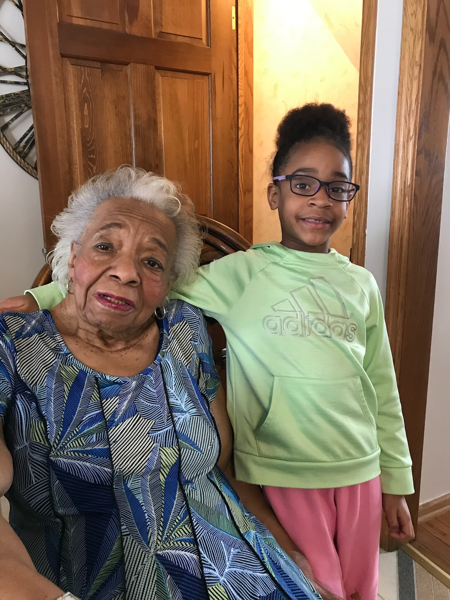 Grandmother and young girl smiling together indoors, standing by a wooden door in a home.