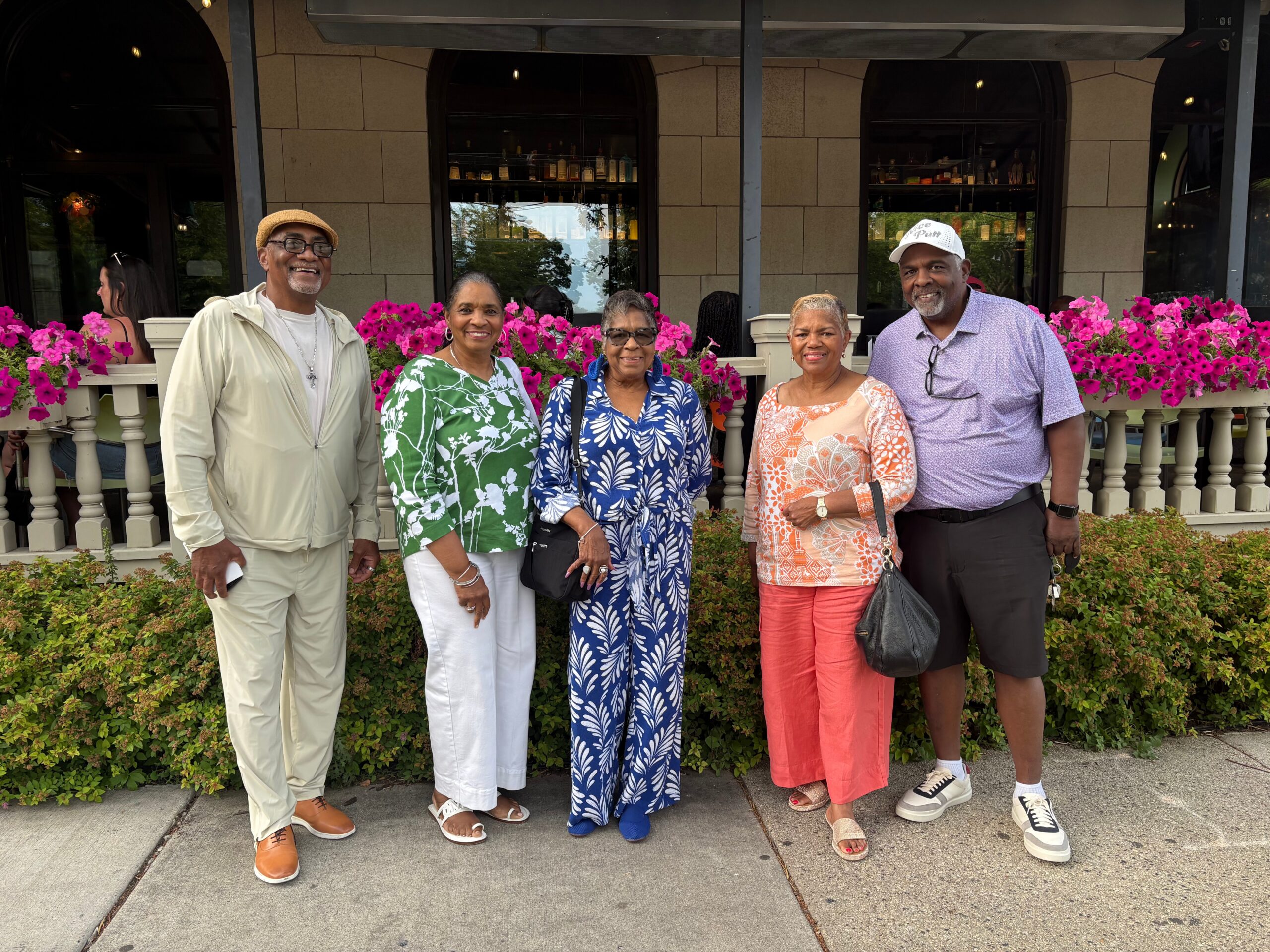 Six ethnically diverse adults posing for a group photo outdoors in front of a building with pink flowering planters, smiling at the camera.