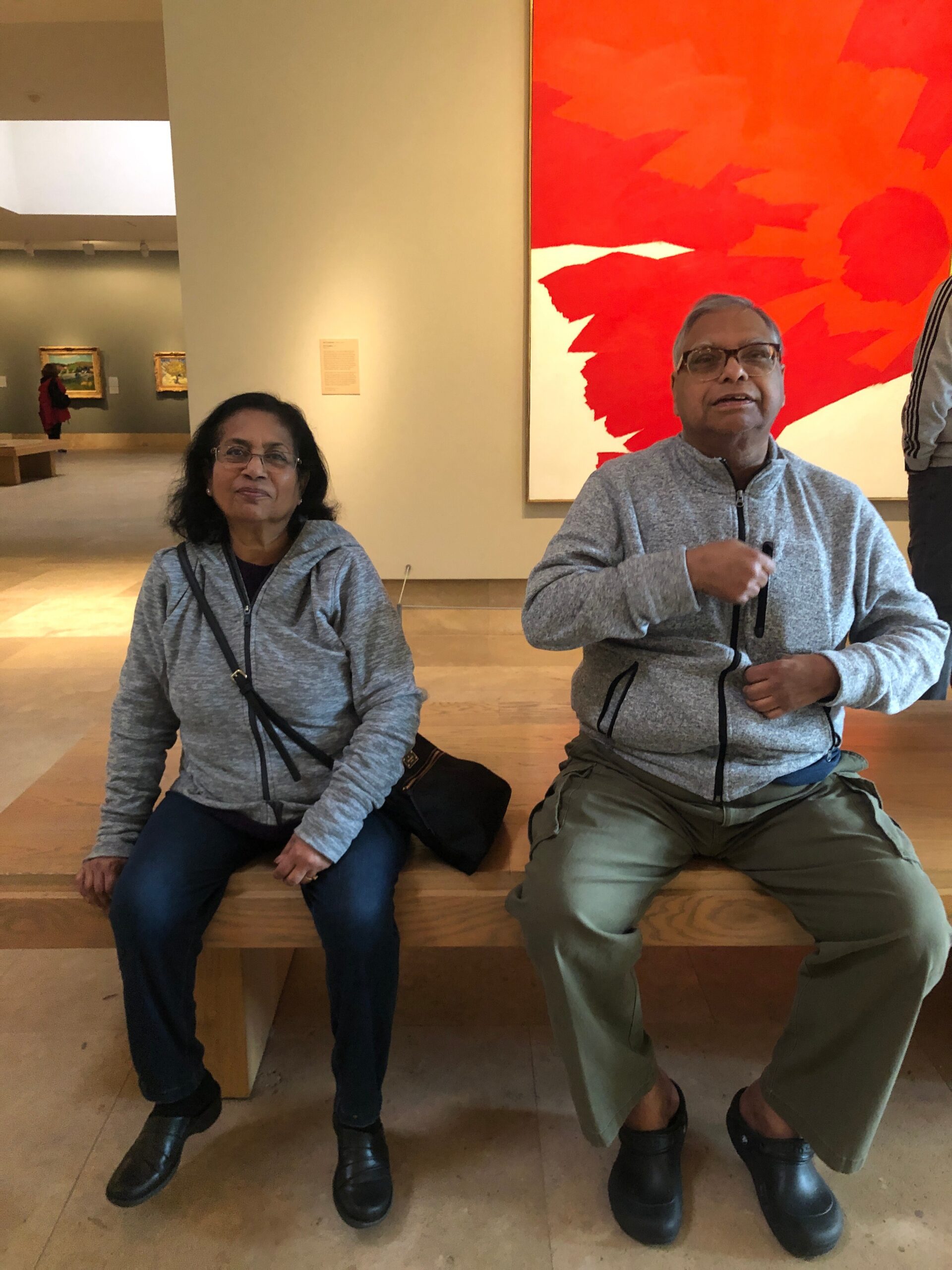 Two seniors sit on a wooden bench in a museum, wearing matching gray zip-up jackets with crossbody bags, in front of a large red abstract painting.