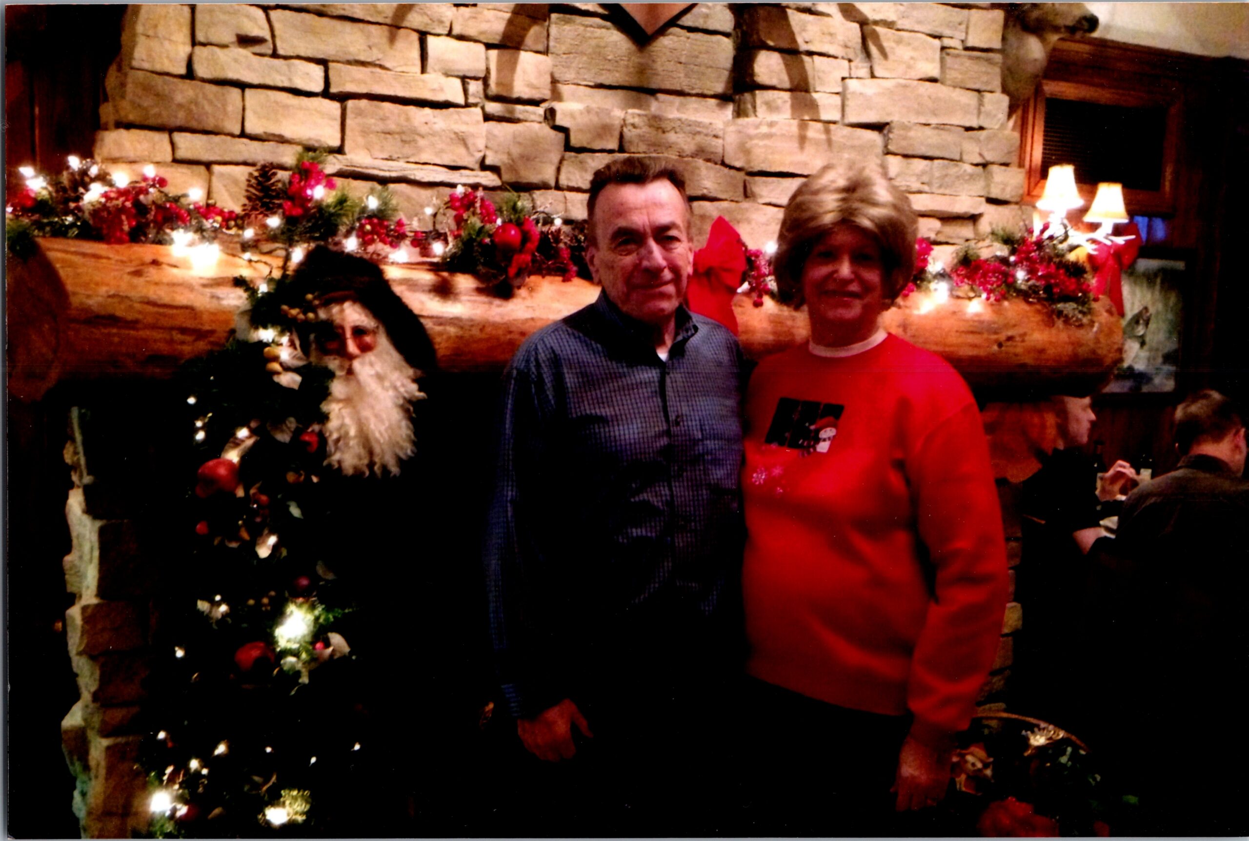 Two adults pose by a stone fireplace adorned with Christmas garland, lights, and red decor; Santa figure visible on the left.