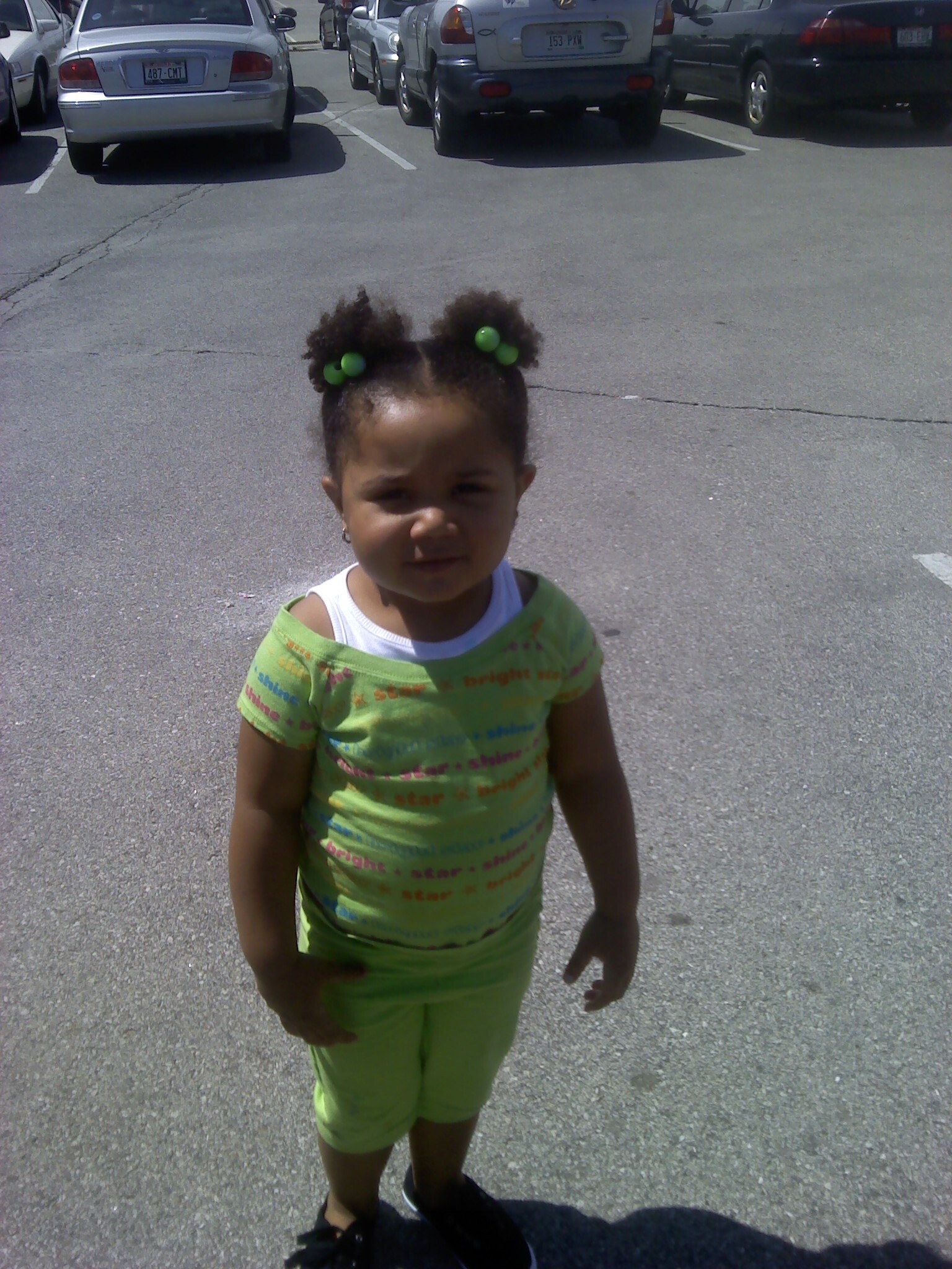 Young girl with green hair accessories stands in a parking lot in front of parked cars, wearing a lime-green outfit.