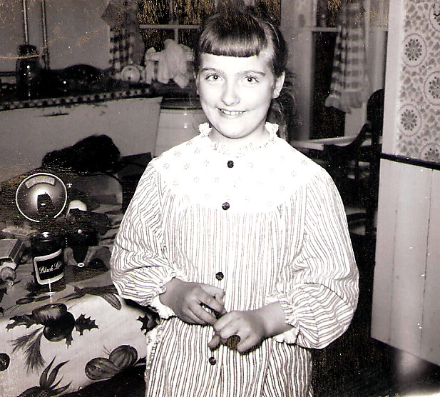 Smiling young girl with bangs in a striped button-front dress, standing in a vintage kitchen.