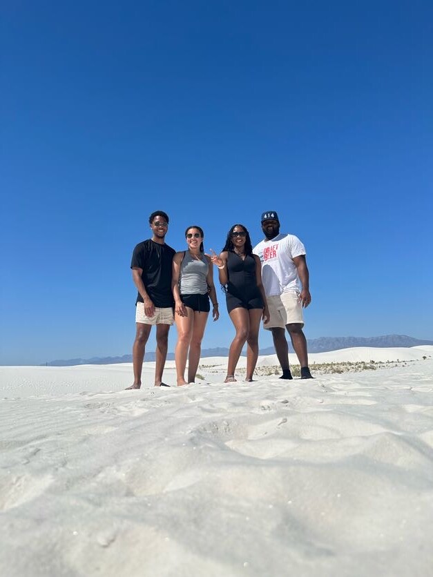Four friends stand together on white sand dunes under a clear blue sky, smiling at the camera with distant mountains on the horizon.