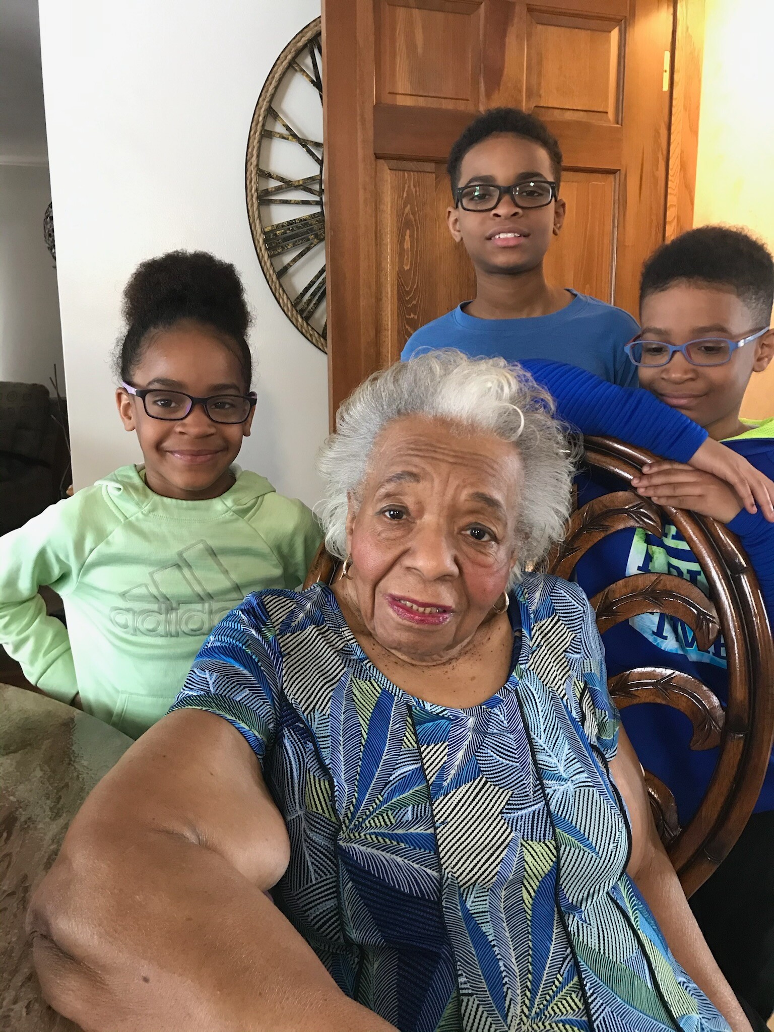 A smiling elderly woman takes a close-up selfie with three children gathered behind her indoors.