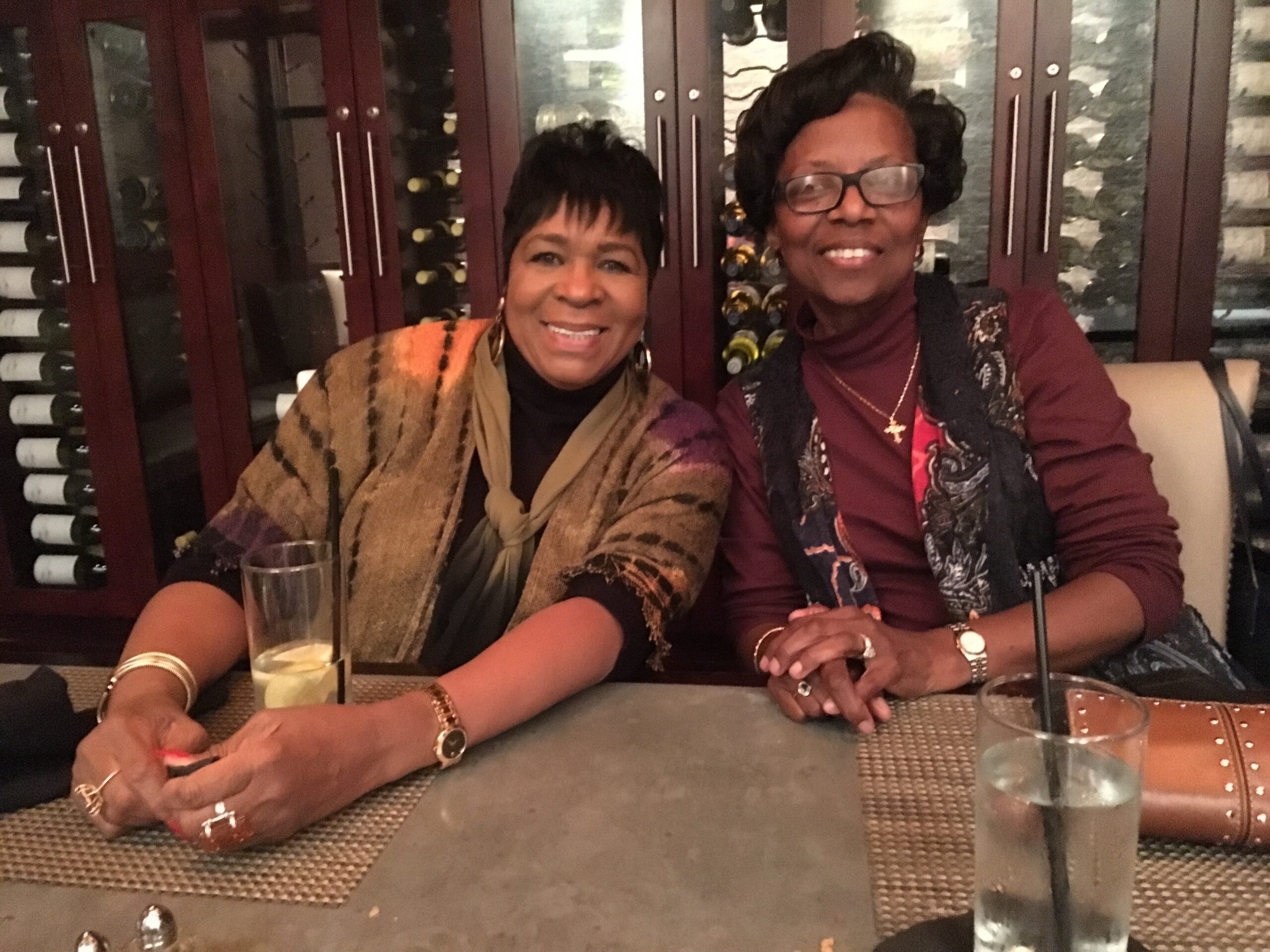 Two smiling women sit side by side at a restaurant table with drinks, posing for a photo in front of a wine rack.