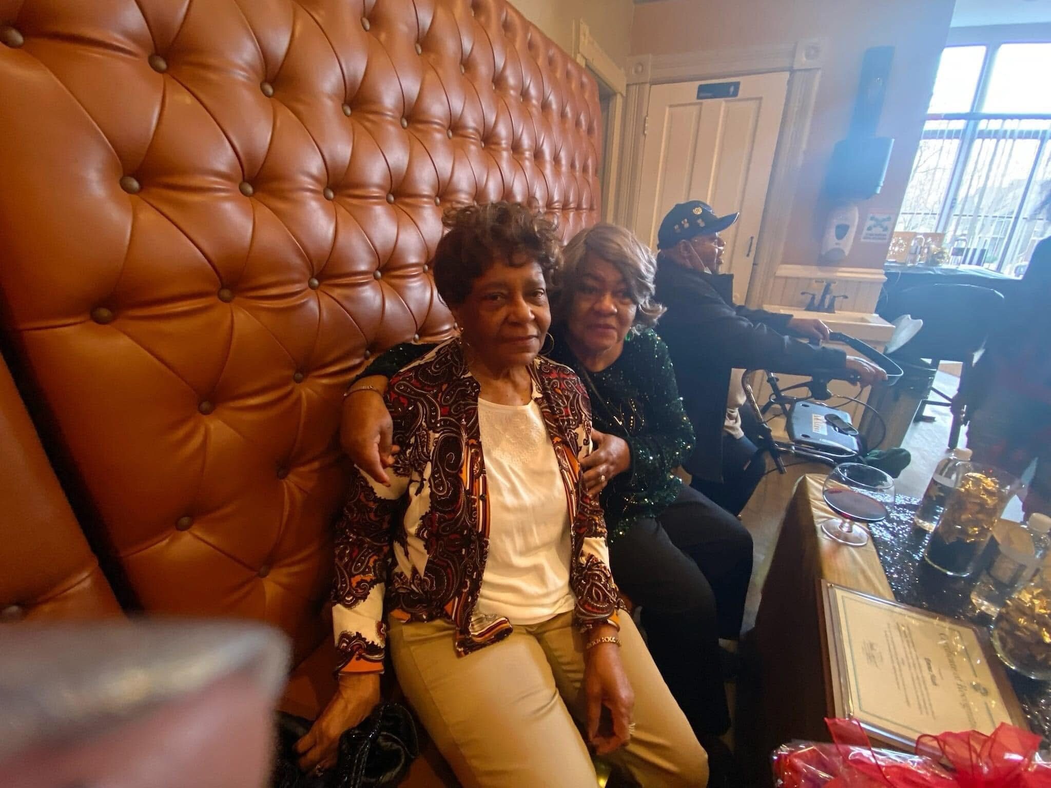 Two elderly women sit together on a large orange tufted bench in a restaurant, smiling at the camera with drinks and menus on the table nearby.