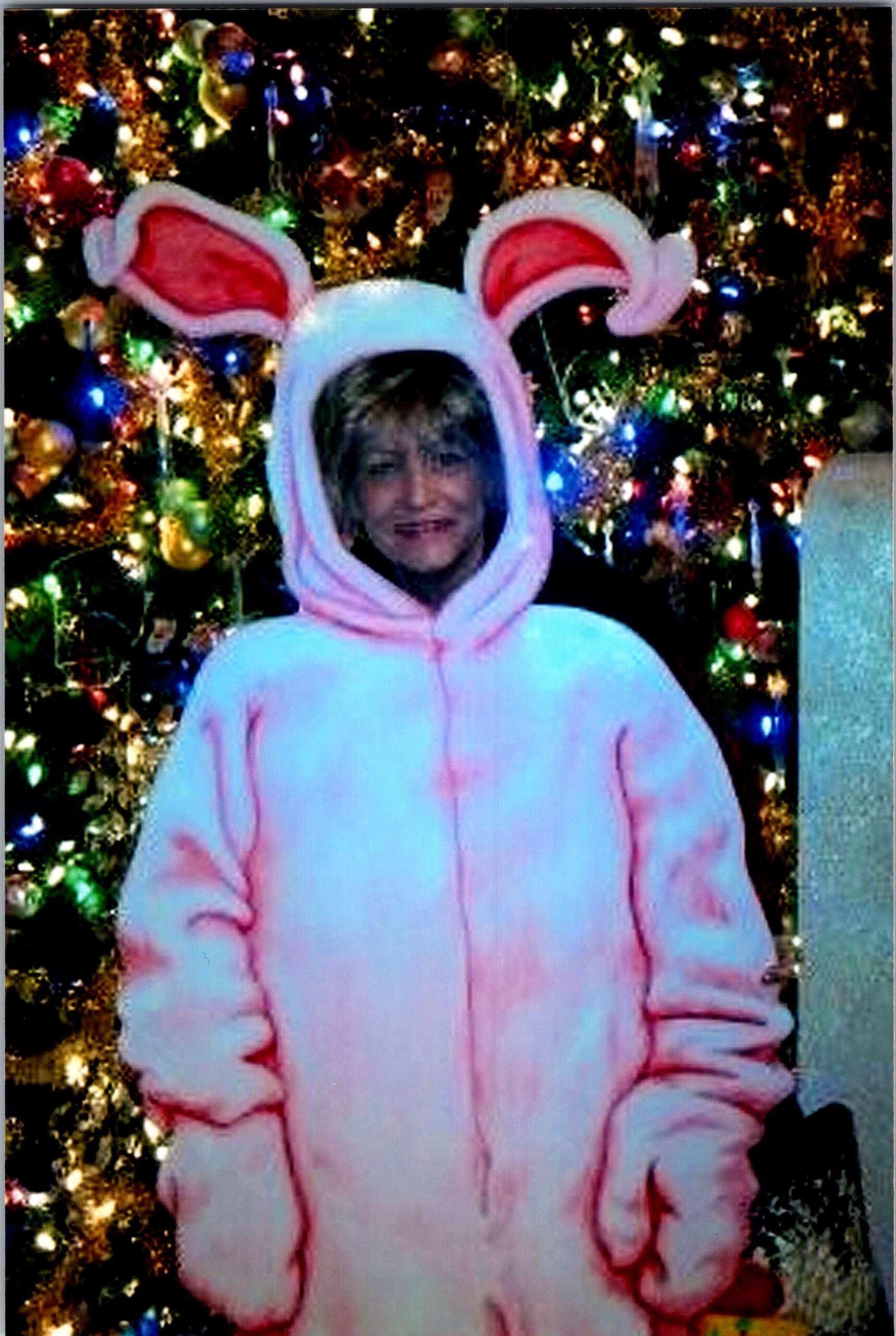Child wearing a pink bunny costume with large ears, smiling in front of a colorful, lit Christmas tree.