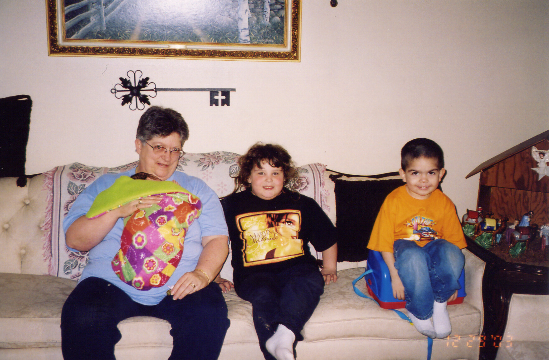 Grandmother sits on a patterned sofa holding a colorful quilt, flanked by two smiling children in a living room.