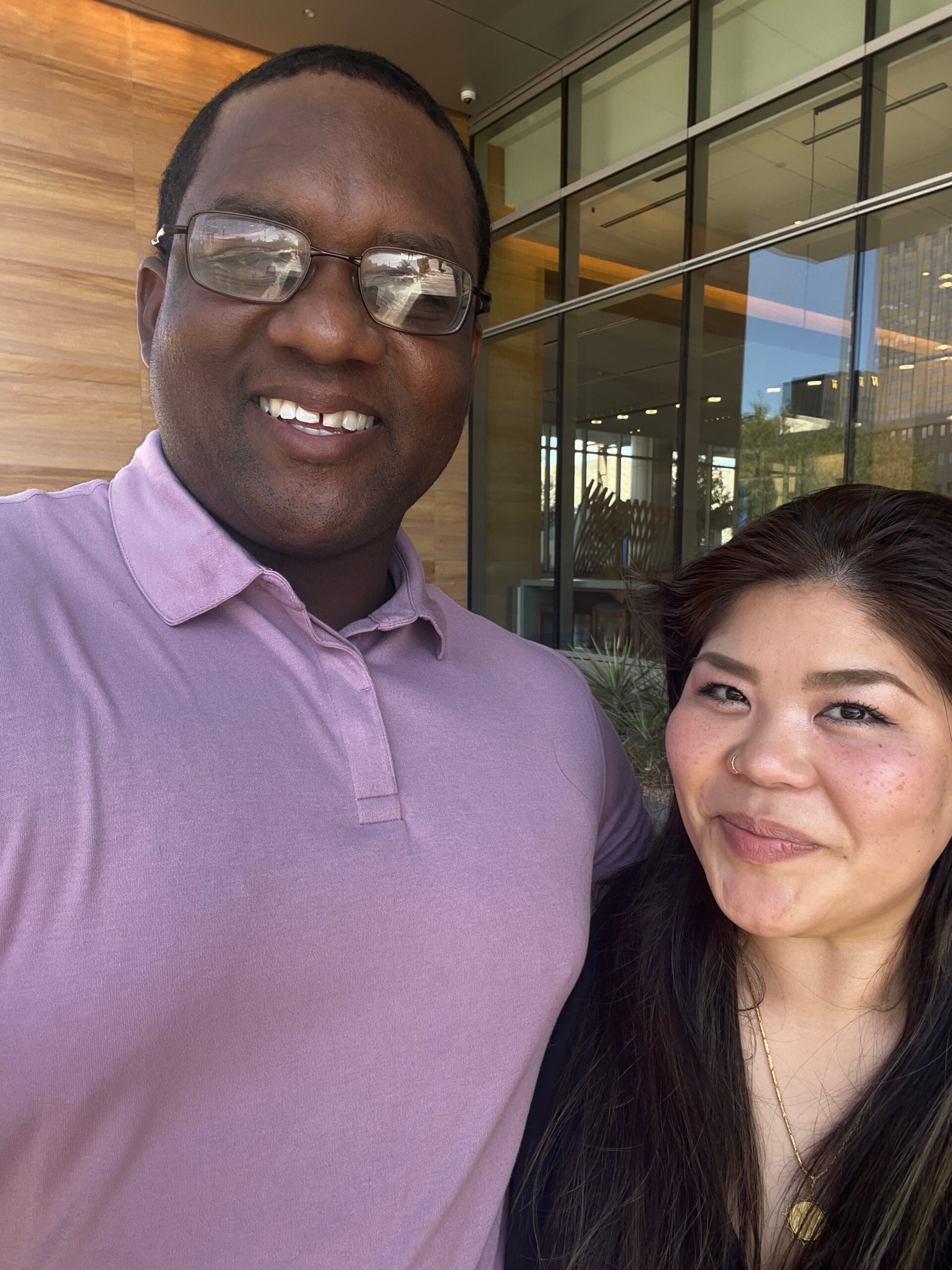 Smiling man wearing glasses and a lavender polo taking a selfie next to a woman in front of a glass-fronted building.