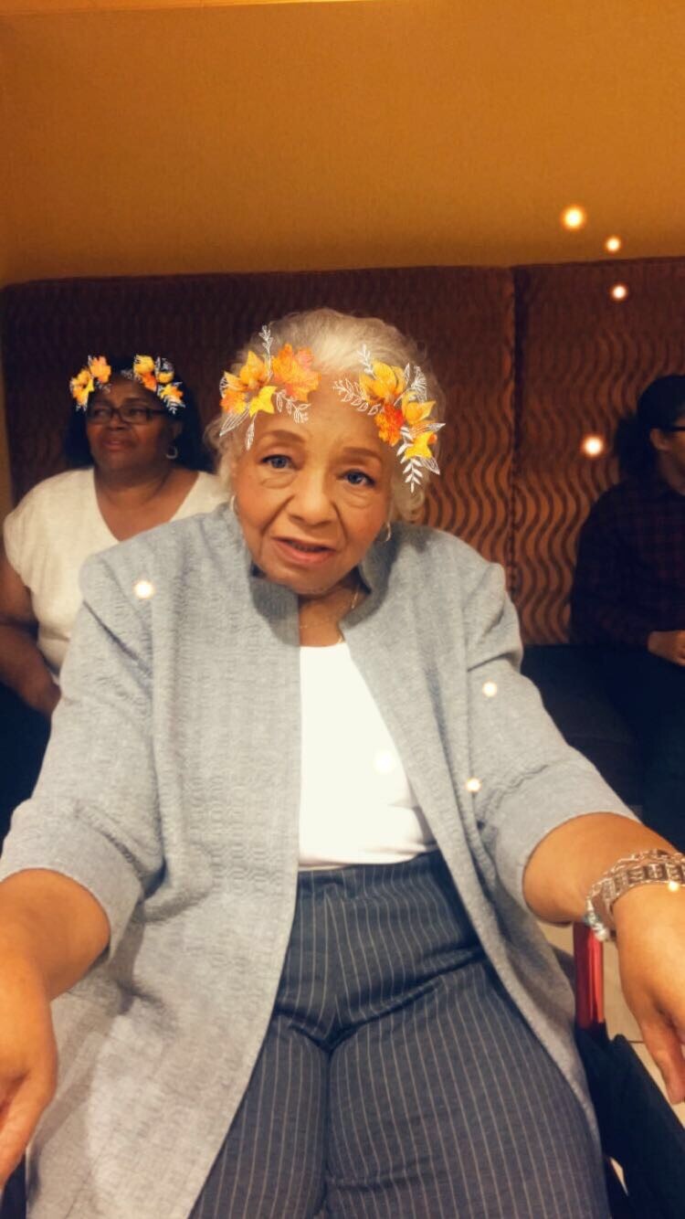Elderly woman seated in a gray cardigan and striped pants, wearing an orange flower crown filter and smiling at the camera.