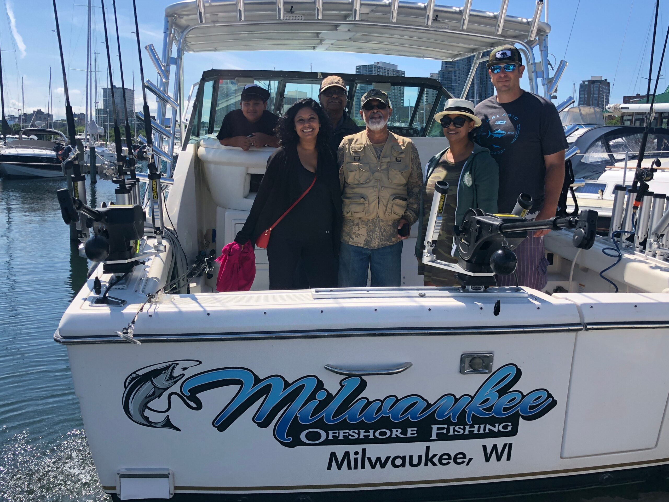 Group of six people posing on a fishing boat with Milwaukee, Wisconsin branding in the foreground, harbor skyline in the background.