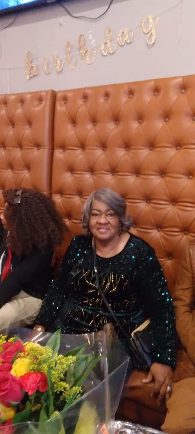 Older woman with gray hair in a sequined dress sitting in a tufted brown booth at a birthday party, with a colorful bouquet in front.