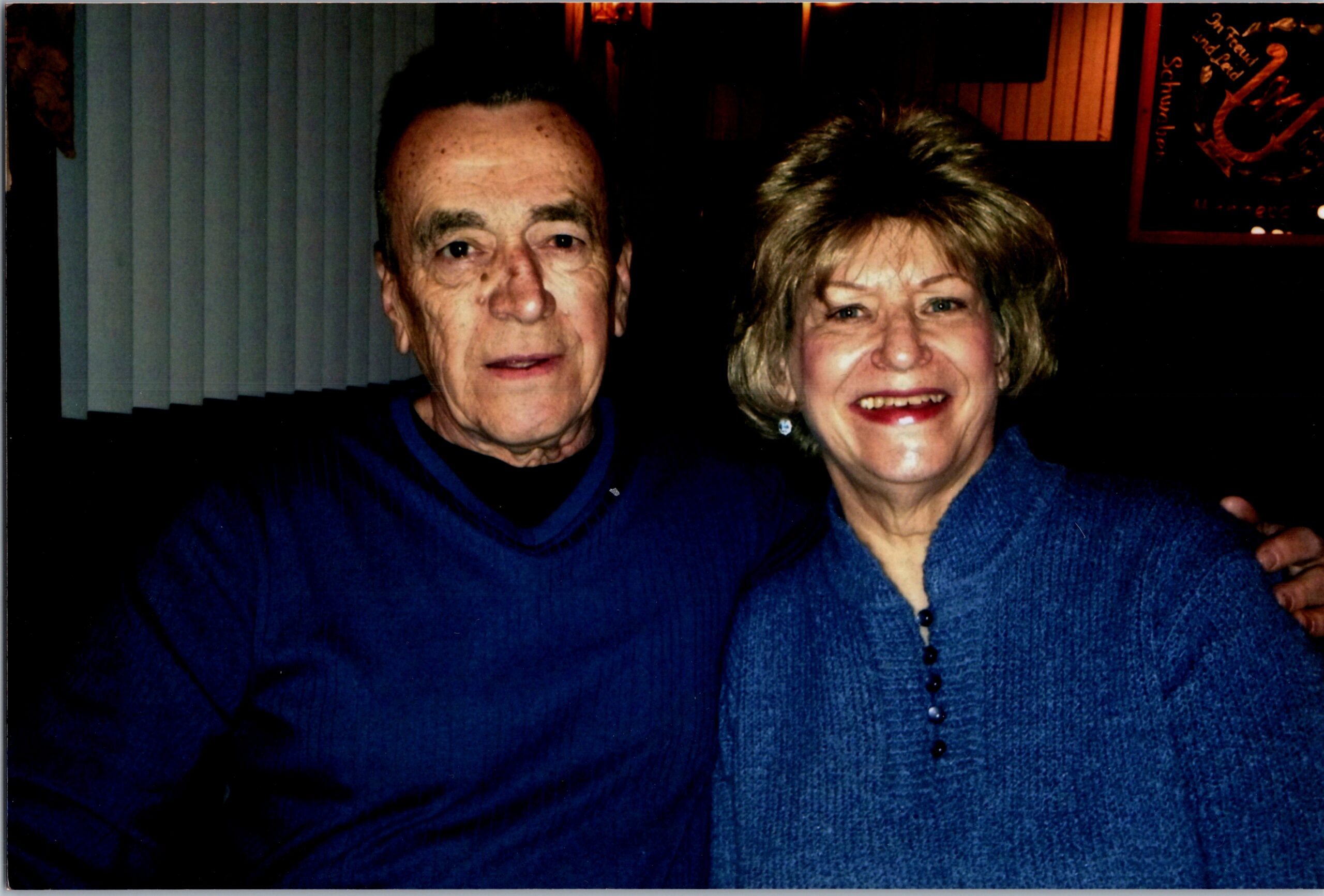 Smiling elderly man and woman posing for a selfie indoors, both wearing blue sweaters; the man on the left and the woman on the right.