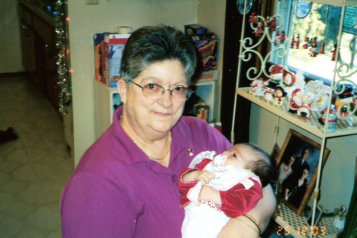 Grandmother in a purple shirt cradling a baby in a red-and-white outfit inside a shop with holiday decor on display.