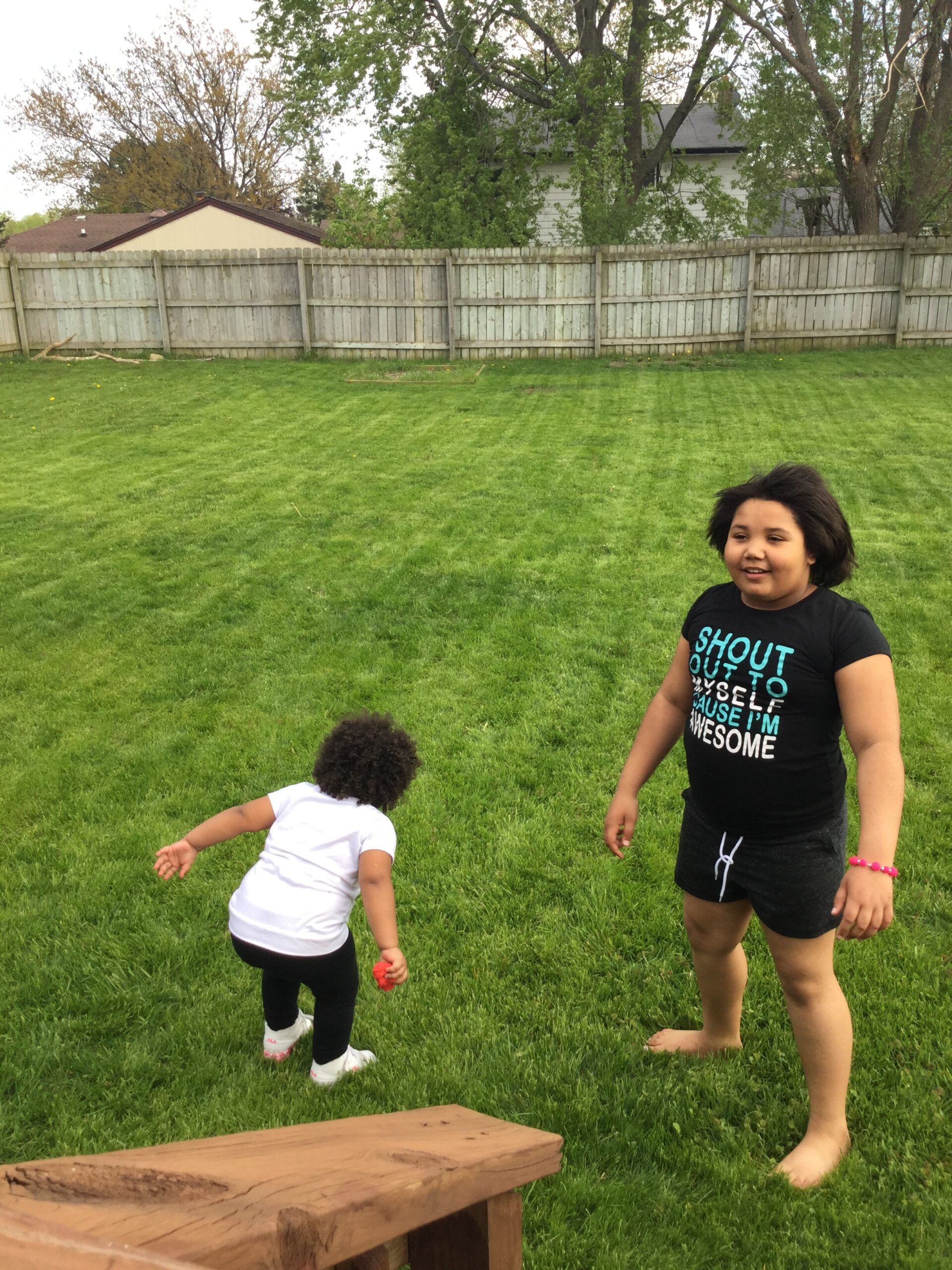 Children playing in a grassy backyard near a wooden fence, one girl standing and smiling while another younger child runs with a small ball or toy nearby.