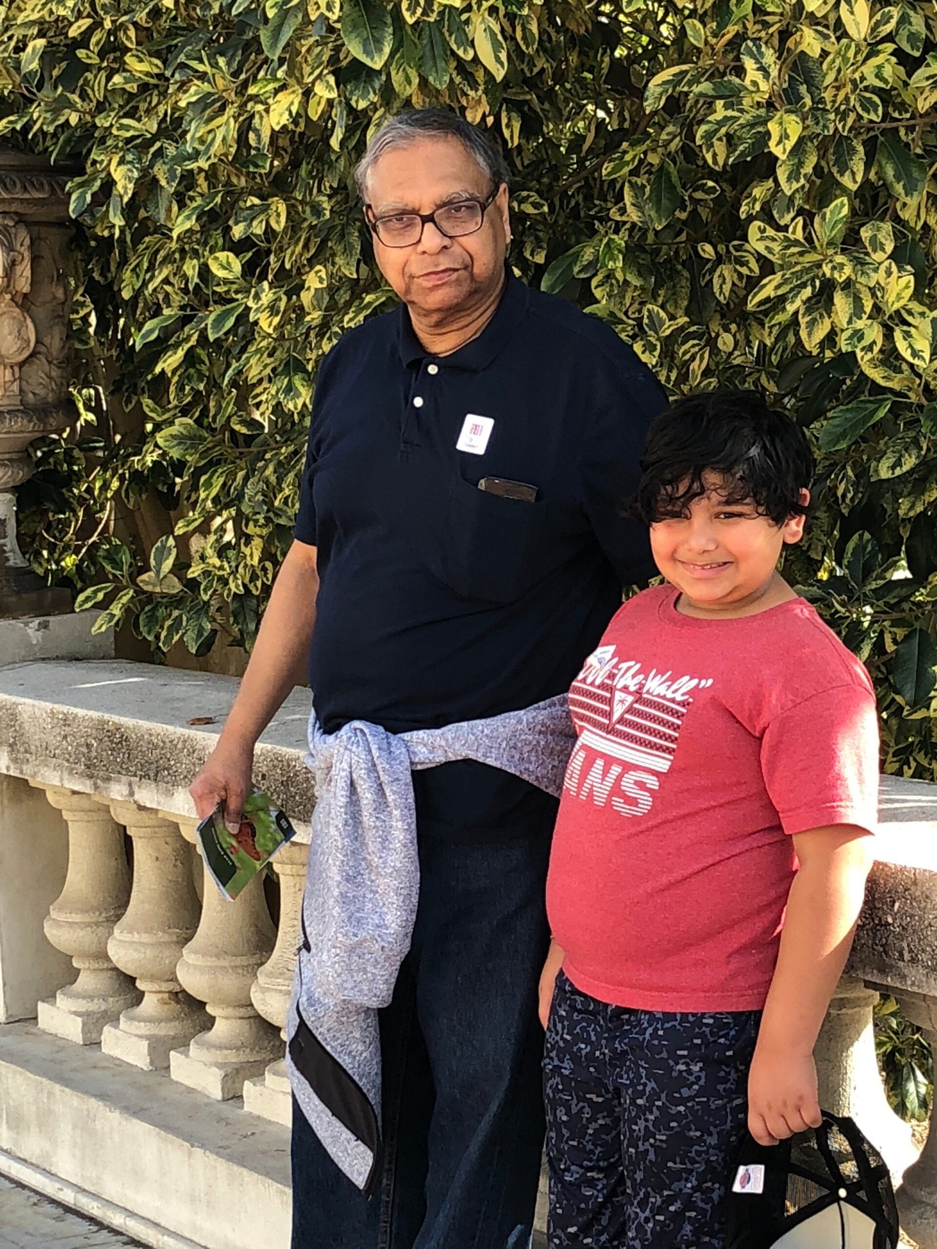 Older man with glasses in a navy polo stands beside a smiling boy in a red shirt, with variegated green-yellow bushes behind them.