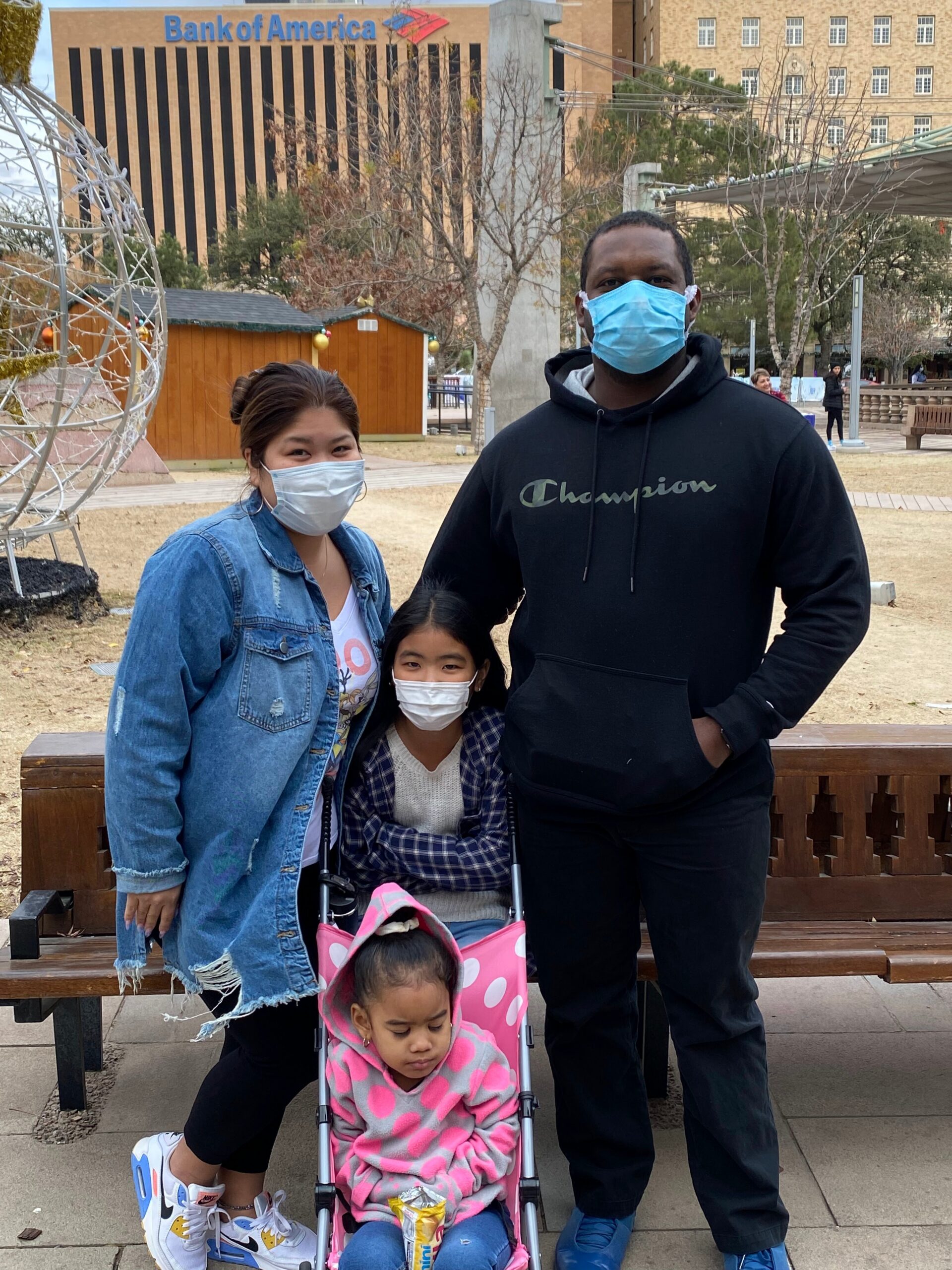 Family of four wearing masks posing in a park; woman in a denim jacket, man in a black hoodie, and two children in a pink stroller, Bank of America building in the background.