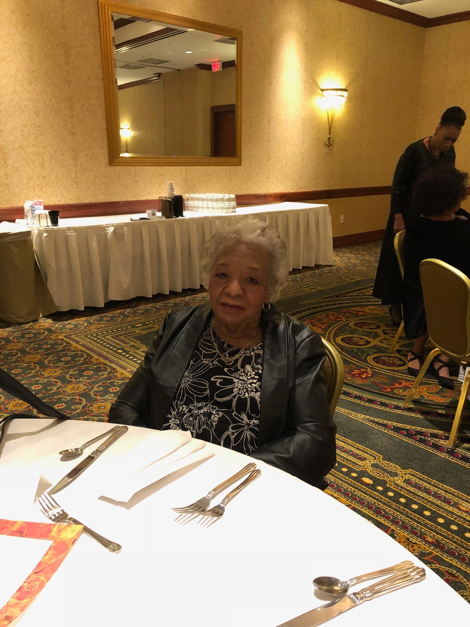 Older woman in a black jacket smiling at a banquet table with silverware in a patterned carpeted room.