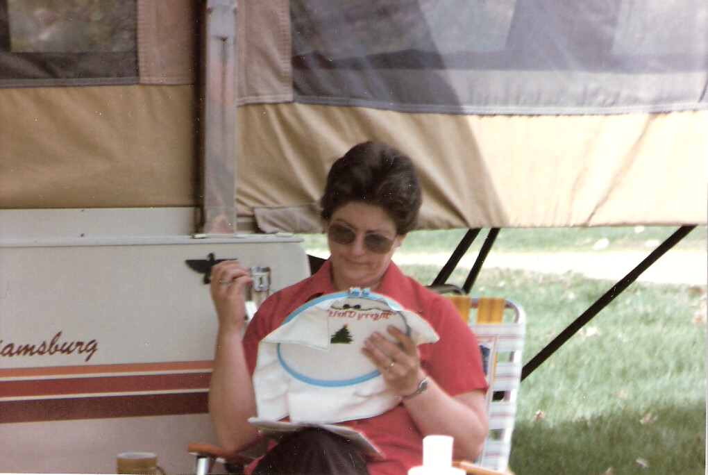 Woman in sunglasses sits in a camping chair beside a popup trailer, threading a needle with embroidery in a hoop showing a pine tree design.