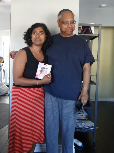 Two adults standing together indoors; the woman in a black top and red striped skirt has her arm around the man and is holding a boxed item facing the camera.