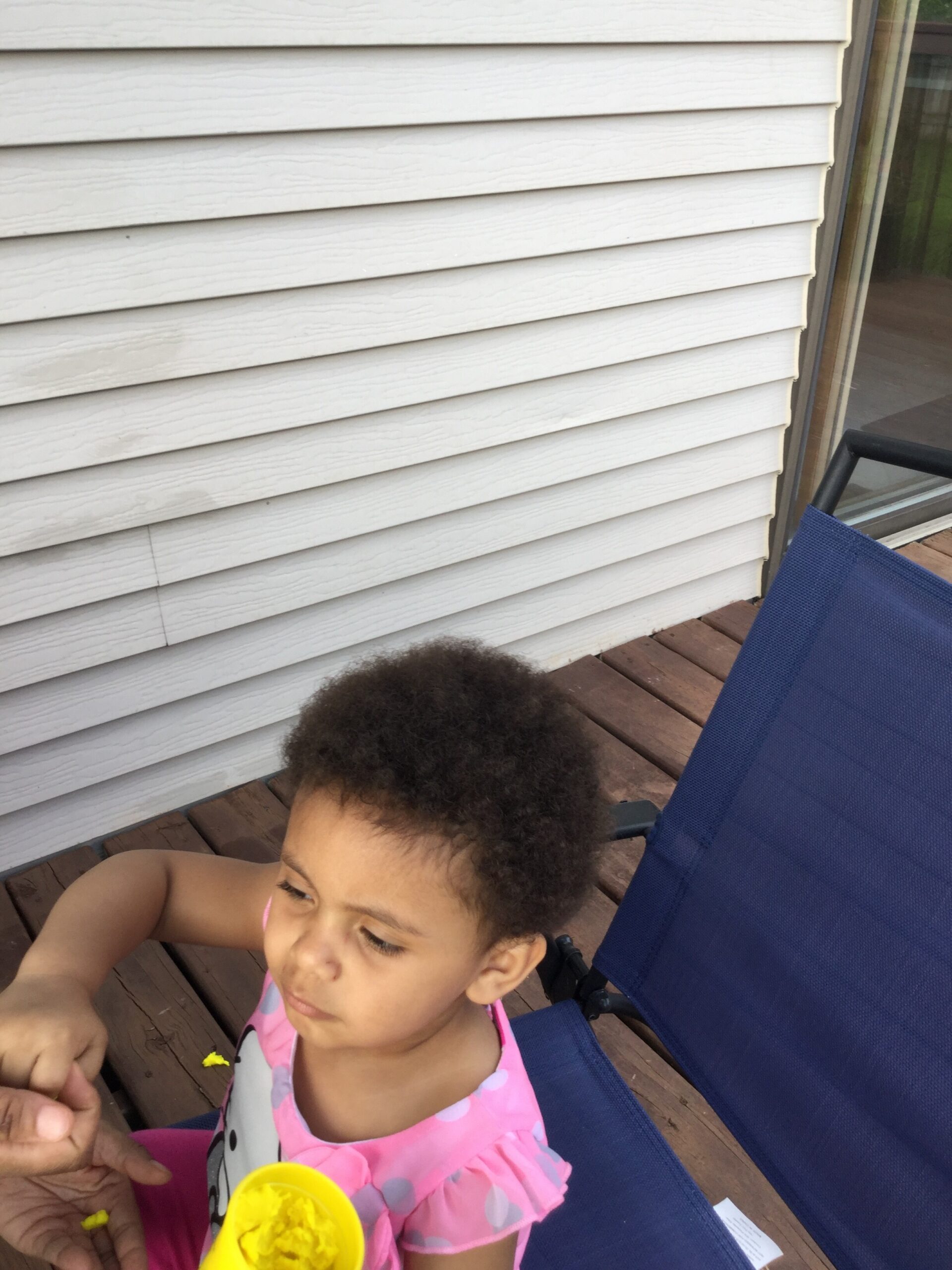 Young child with curly hair sits in a blue outdoor chair on a wooden deck, wearing a pink swimsuit and eating from a yellow cup outdoors.